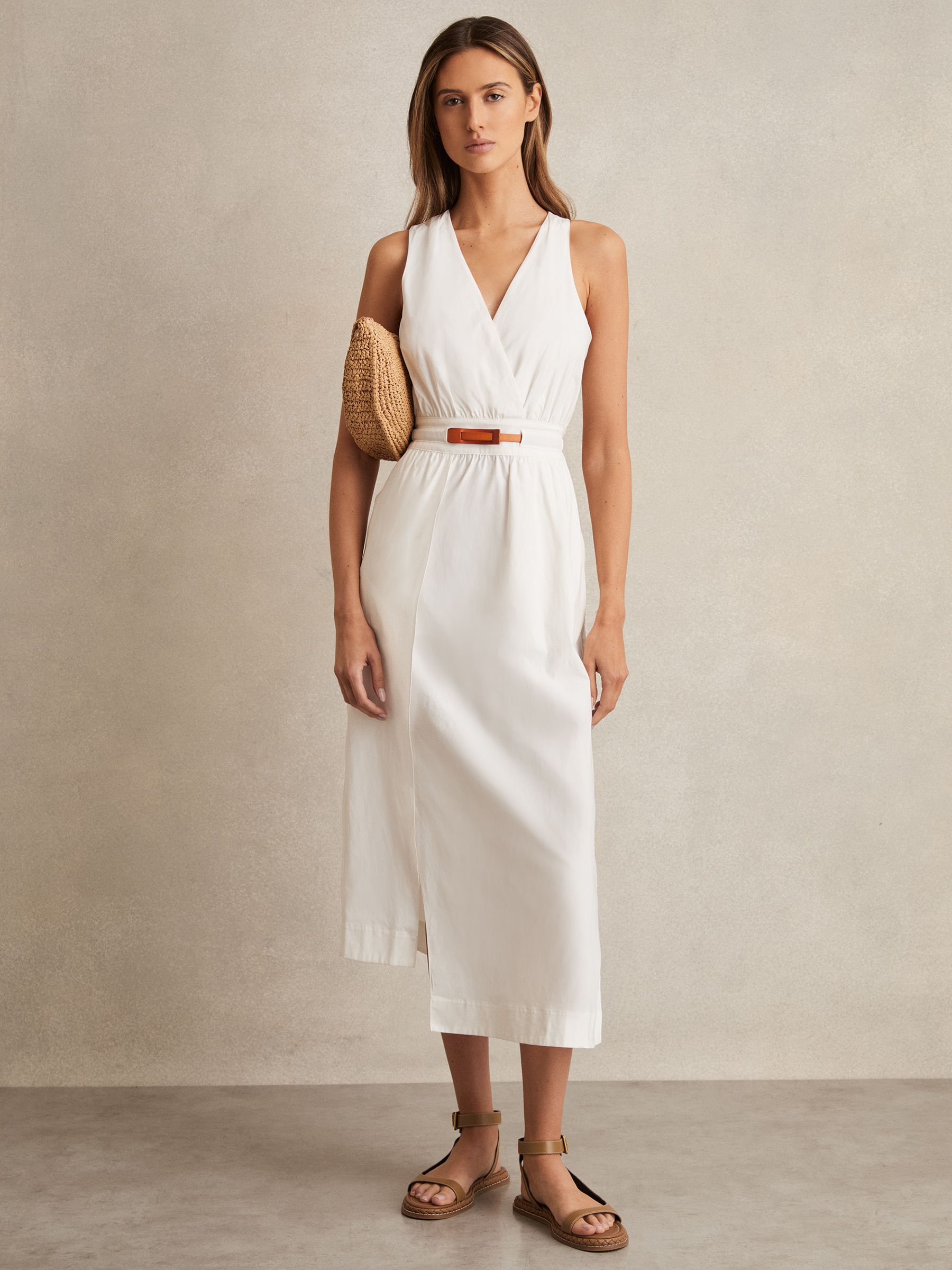 A woman wearing an ivory midi dress with a V-neckline, split skirt, asymmetric hemline, and a contrasting orange leather belt, standing against a neutral background.