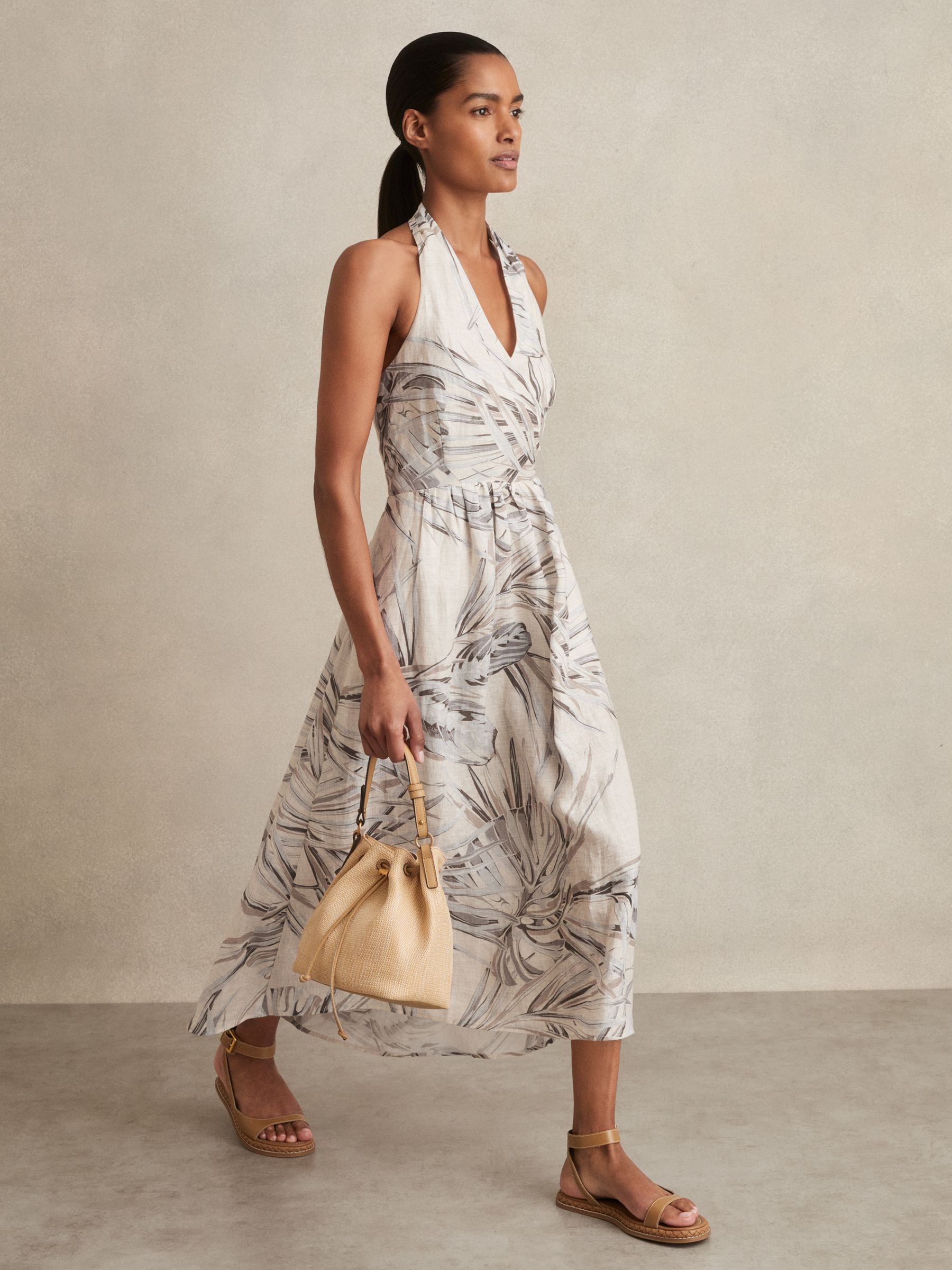 A woman wearing a sleeveless, neutral-colored linen midi dress with a palm print, halterneck with buttons, and a wrap V-neckline, holding a beige handbag and wearing beige sandals.