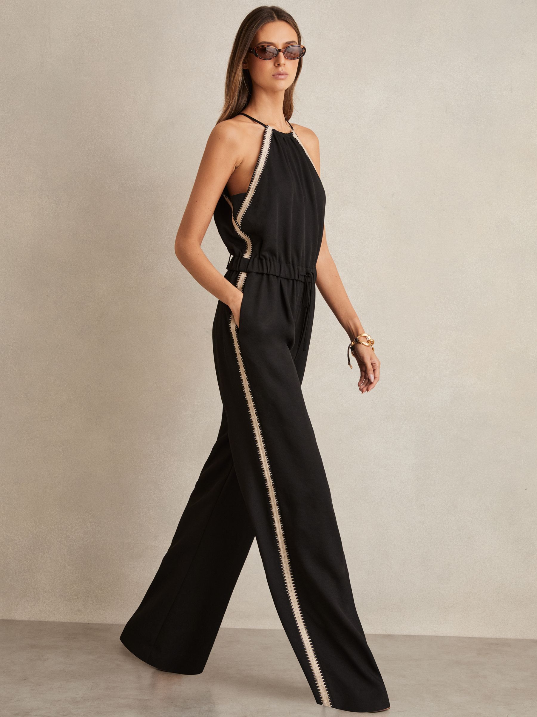 A woman wearing a black halterneck jumpsuit with embroidery details, wide-leg design, and a self-tie halterneck.