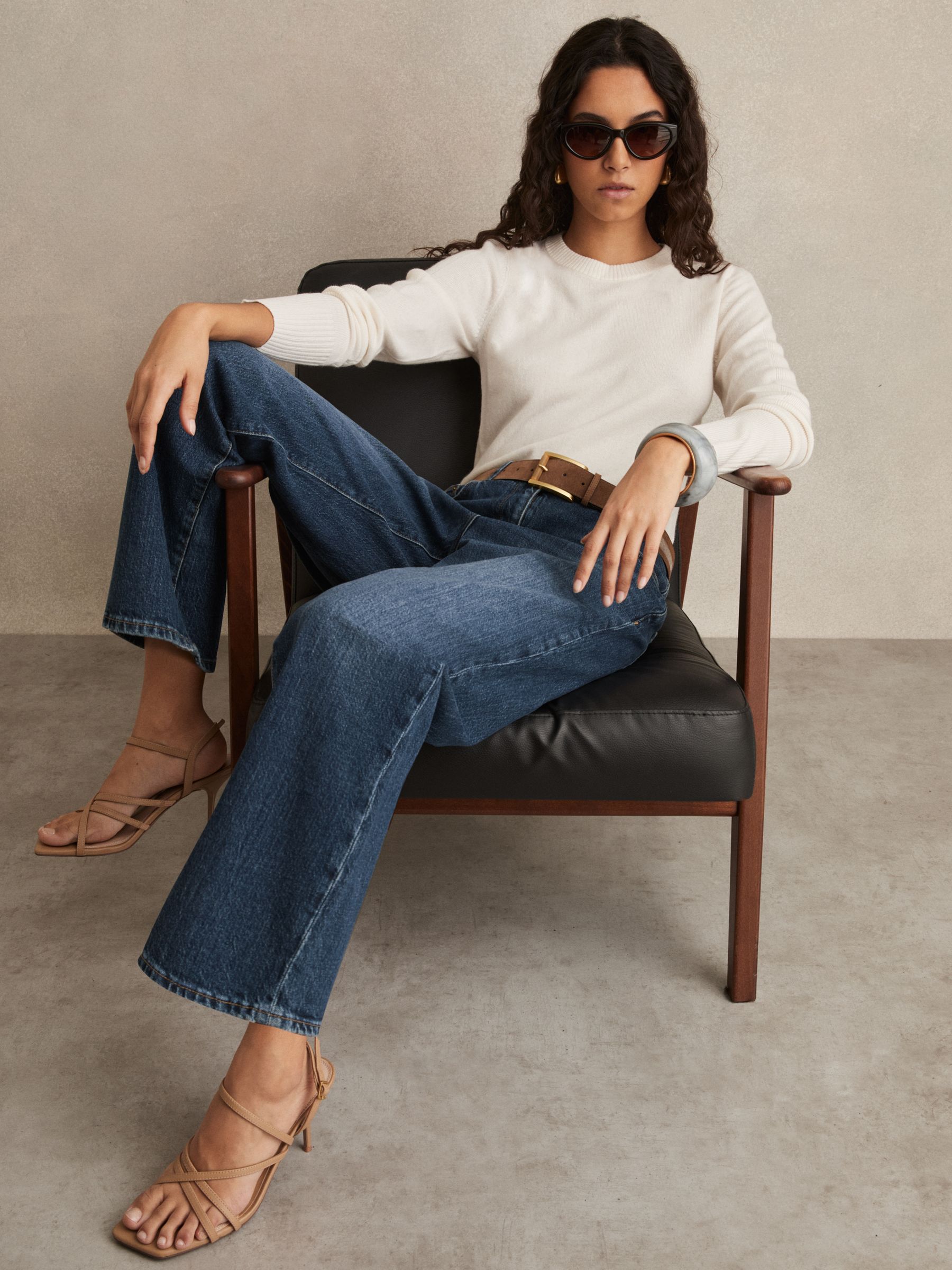 A woman sitting in a chair wearing mid blue straight leg jeans, a white sweater, beige high-heeled sandals, sunglasses, and gold earrings.