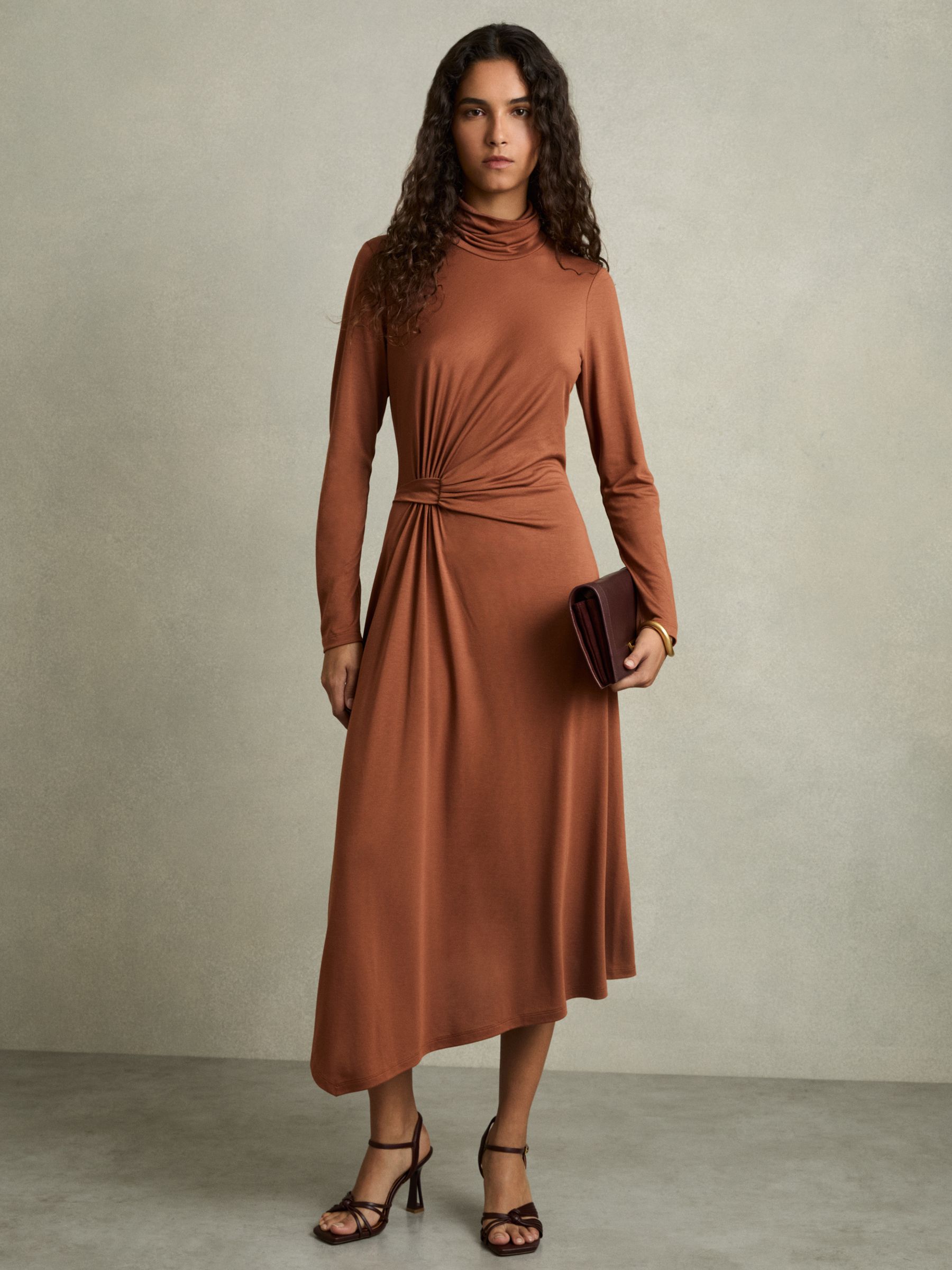 A woman wearing a camel-colored midi dress with asymmetric draping, a roll neck, and a side slit, paired with high-heeled sandals and holding a clutch.