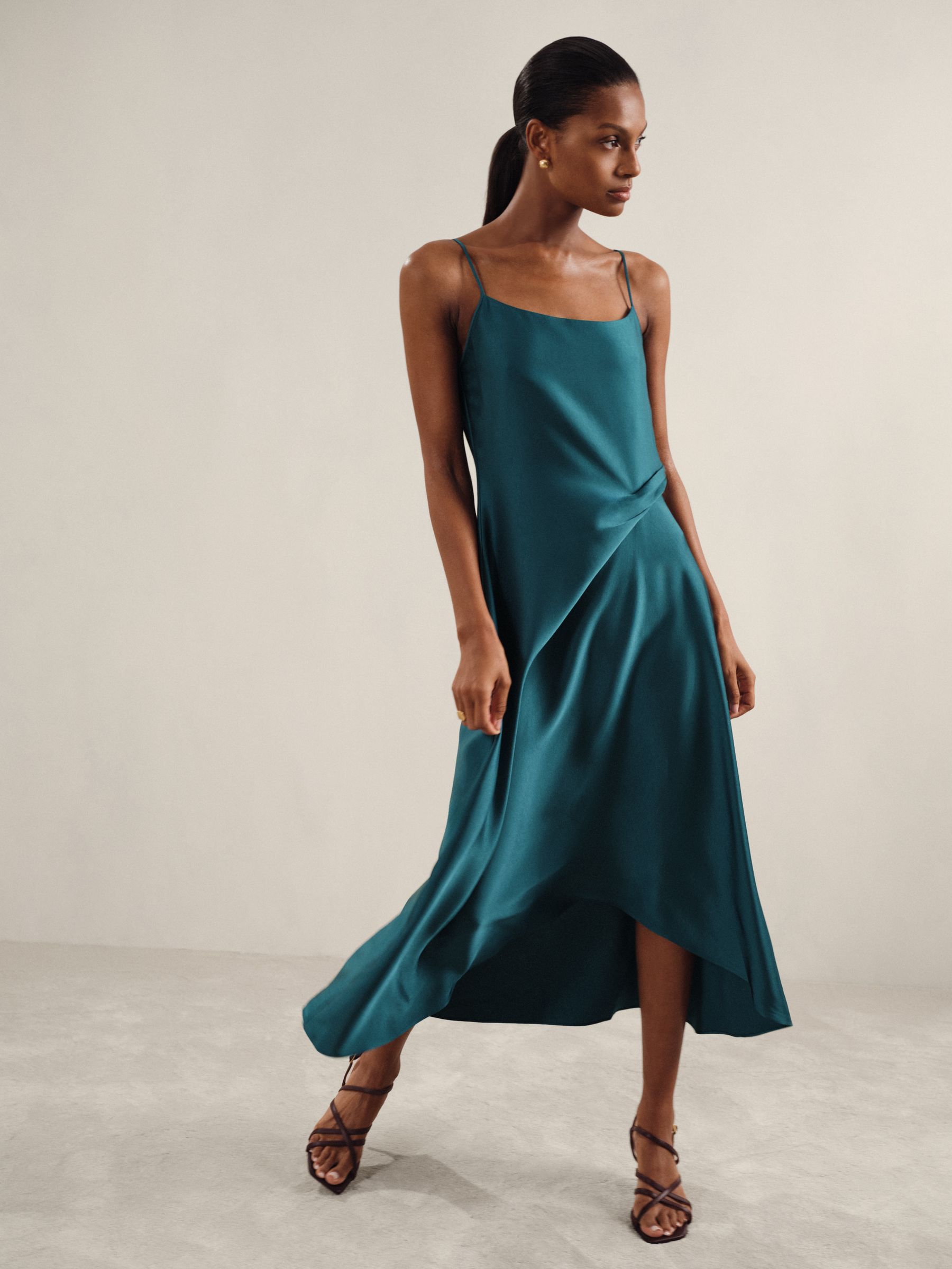 A woman wearing a teal satin slip dress with thin straps, drape detail at the waist, and an asymmetric midi-length hem.