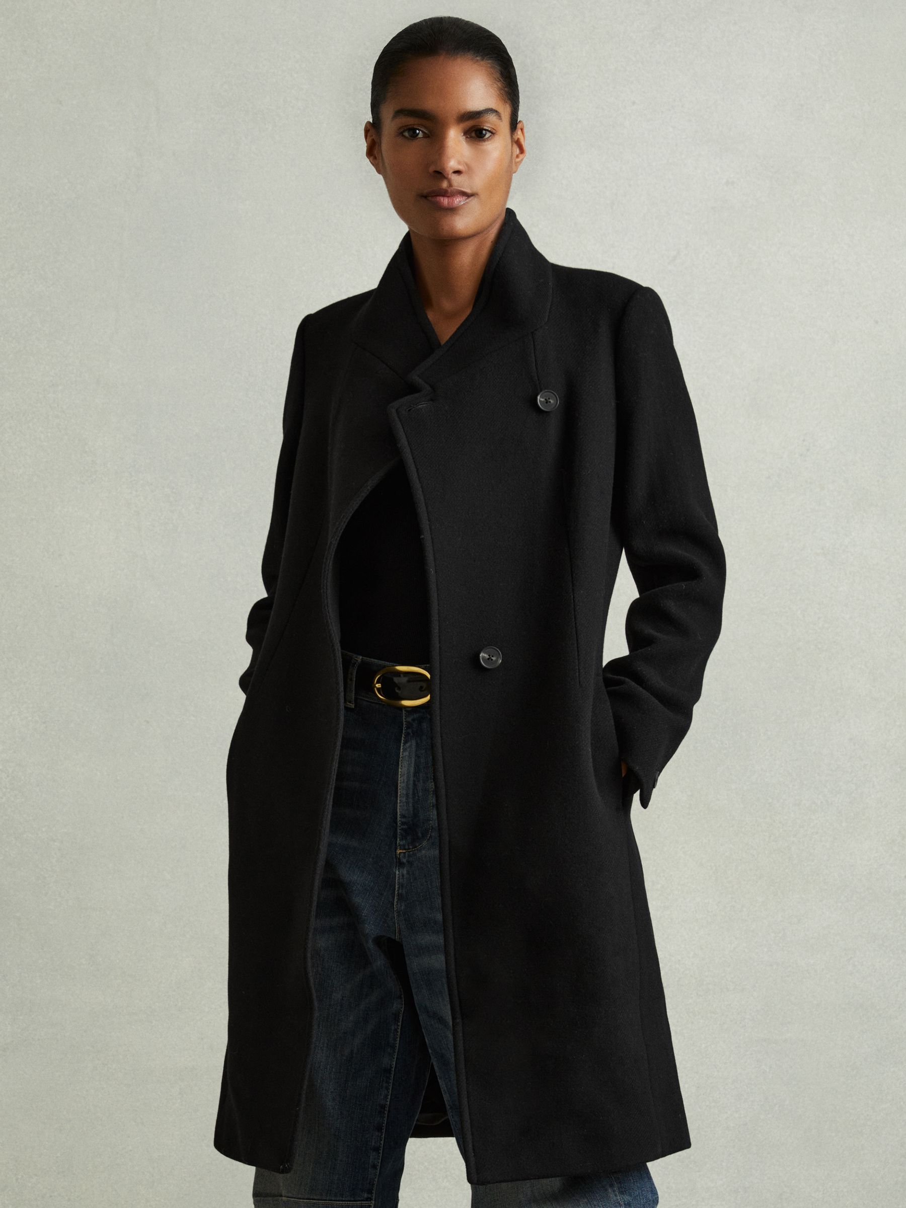 A woman wearing a black longline wool-blend coat with a funnel neckline, double-breasted front, flap pockets, and lining.