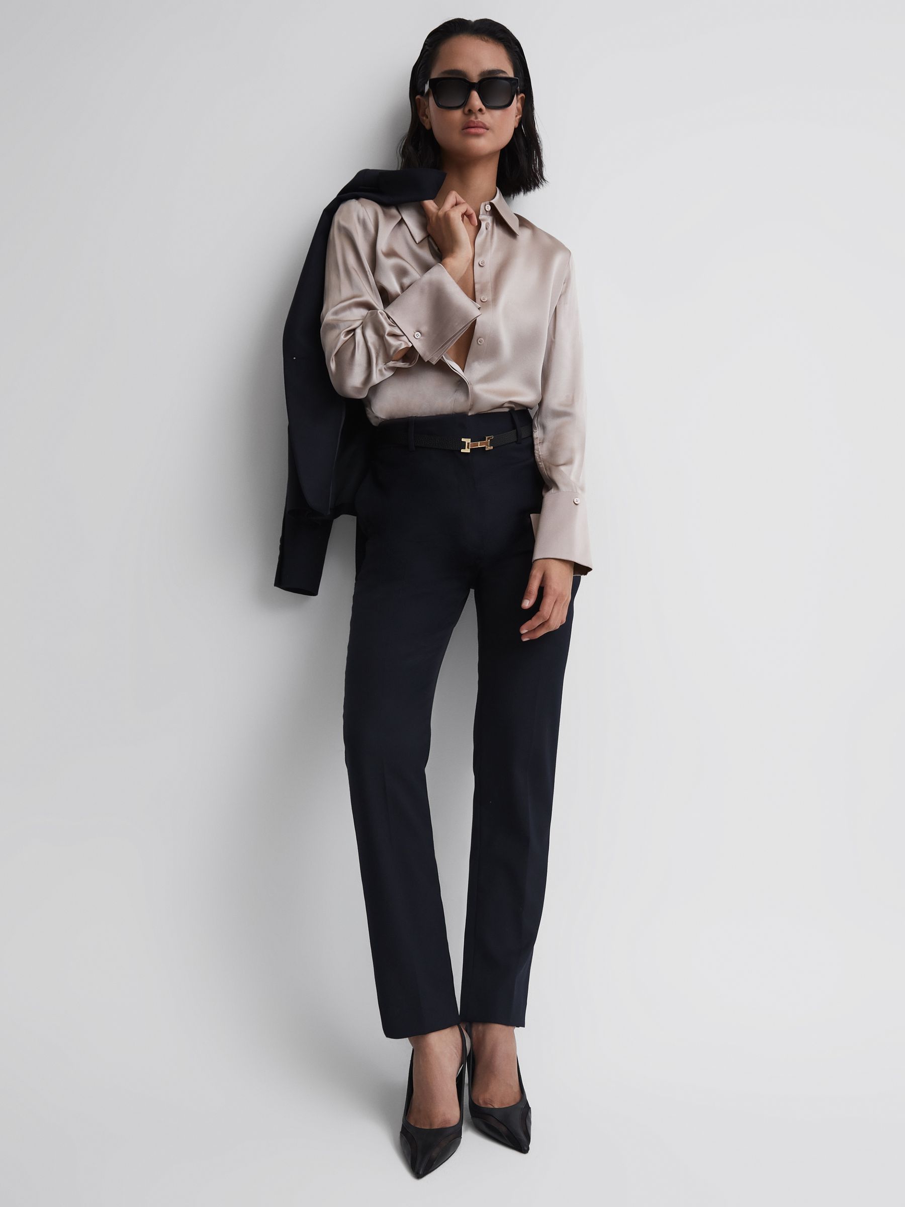A woman wearing navy tapered suit trousers with a beige silk blouse, black heels, and sunglasses.