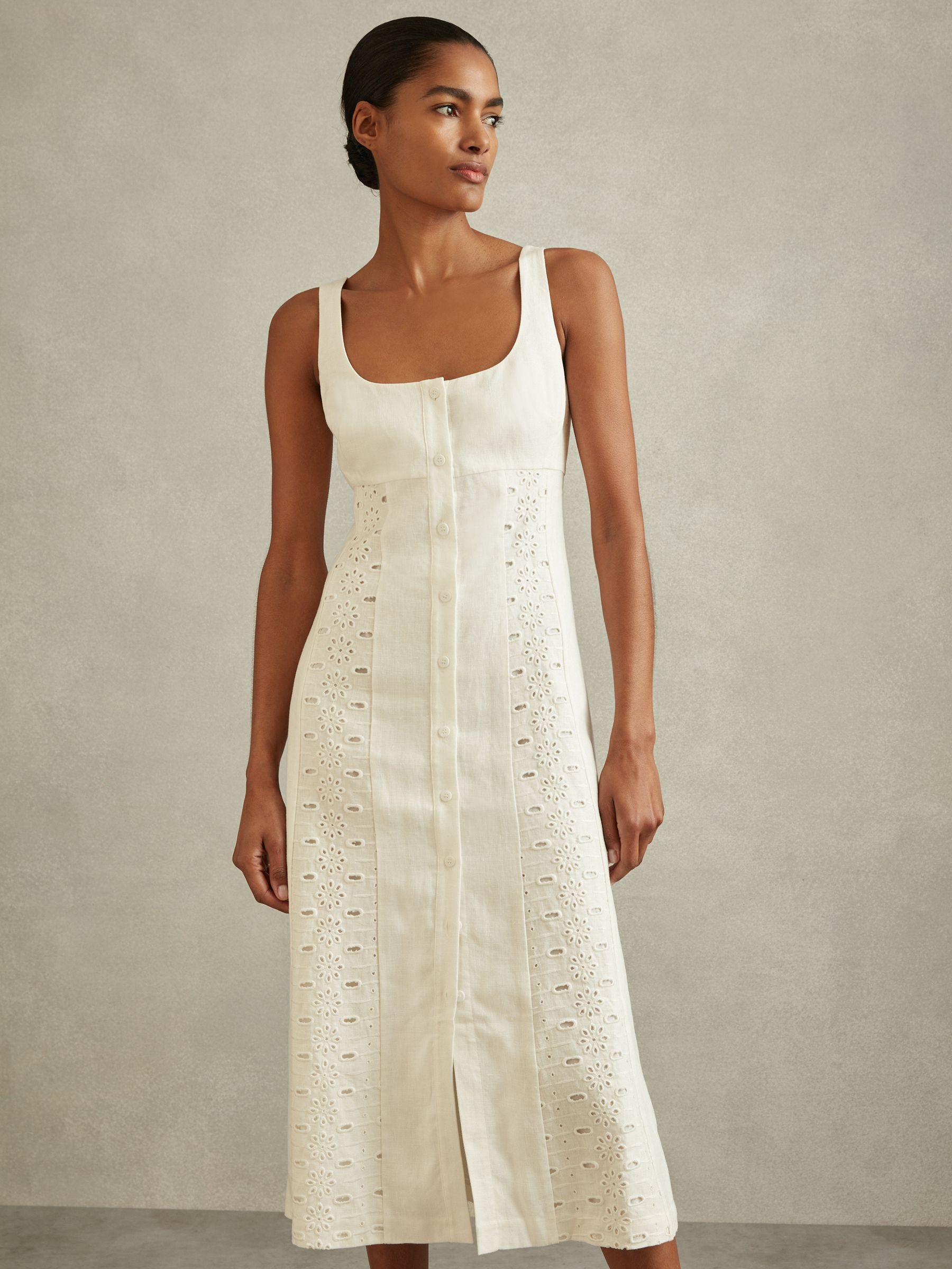 A woman wearing an ivory linen midi dress with broderie detailing, adjustable shoulder straps, and a button-front closure.