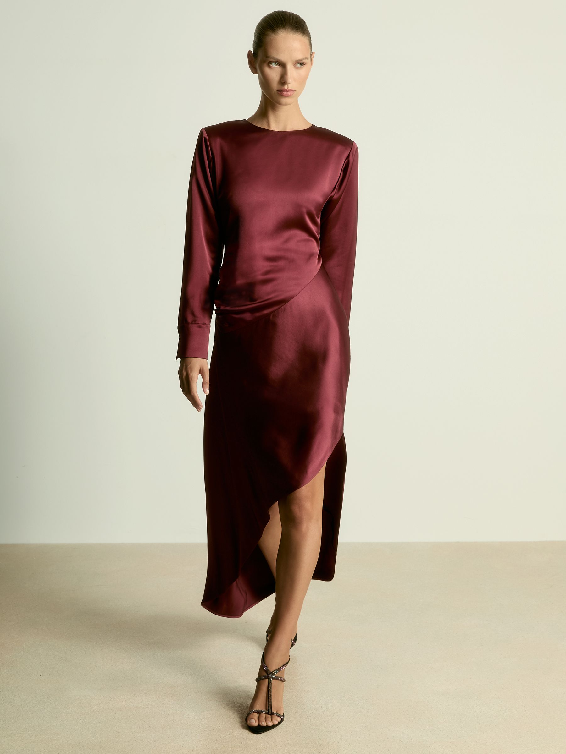 A woman wearing a burgundy satin maxi dress with asymmetric draping, curved hem with side slit, and buttoned cuffs.