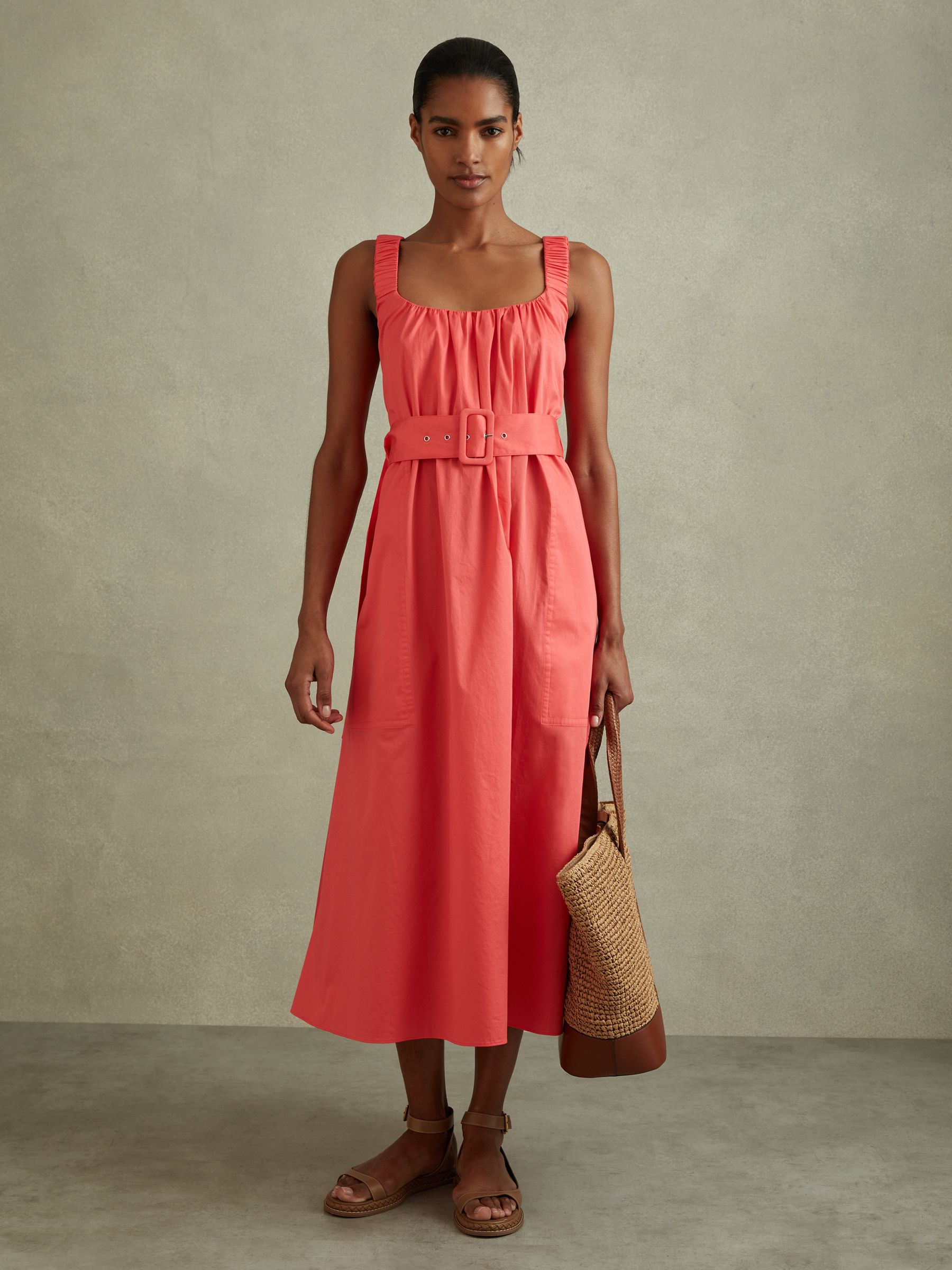 A woman wearing a coral midi dress with pleats at the neckline, ruched shoulder straps crossing at the back, and a wide belt at the waist.