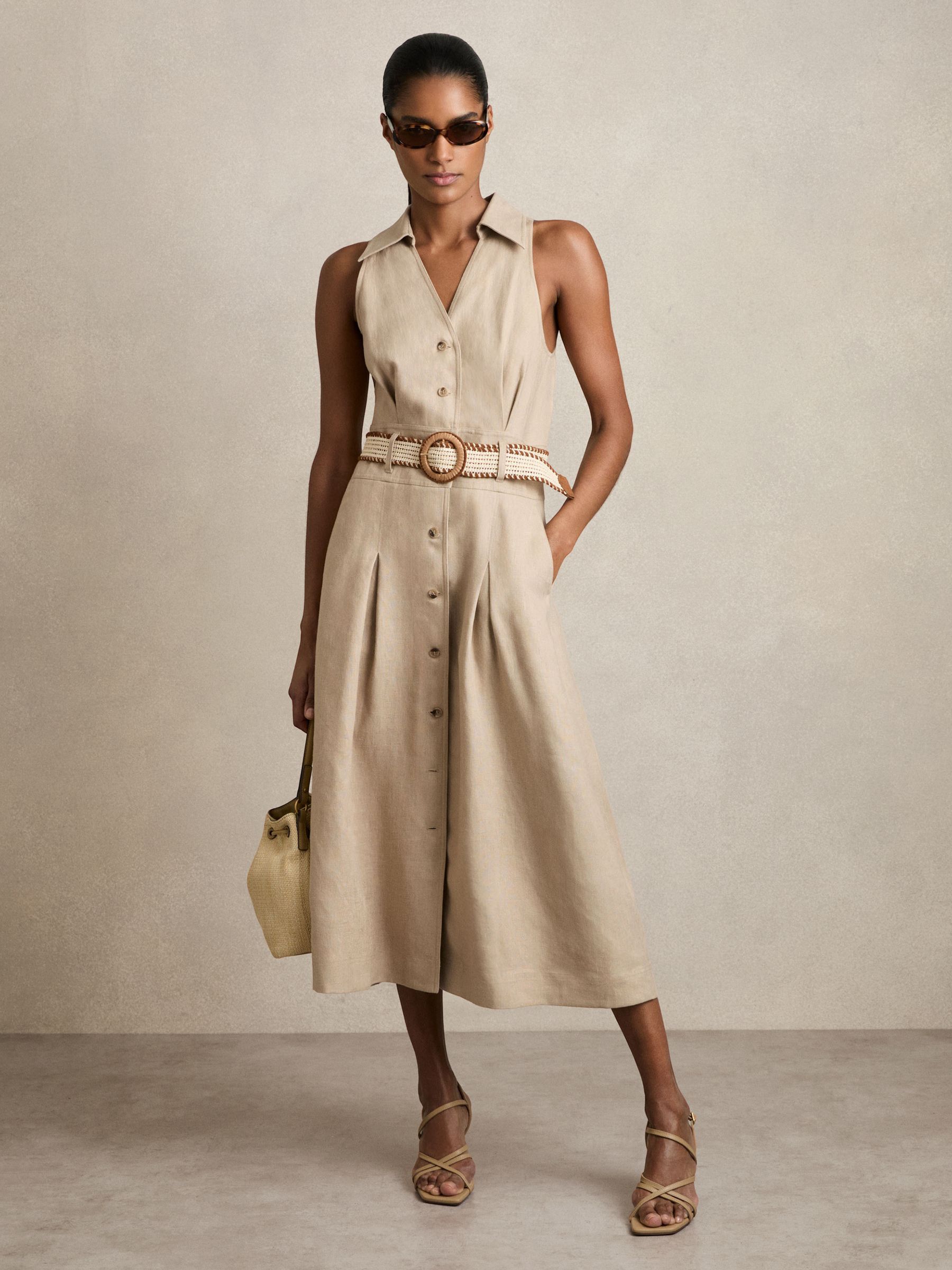 A woman wearing a beige sleeveless linen midi dress with a V-neckline, collar, front buttons, pleated skirt, and a raffia woven belt with suede trim.