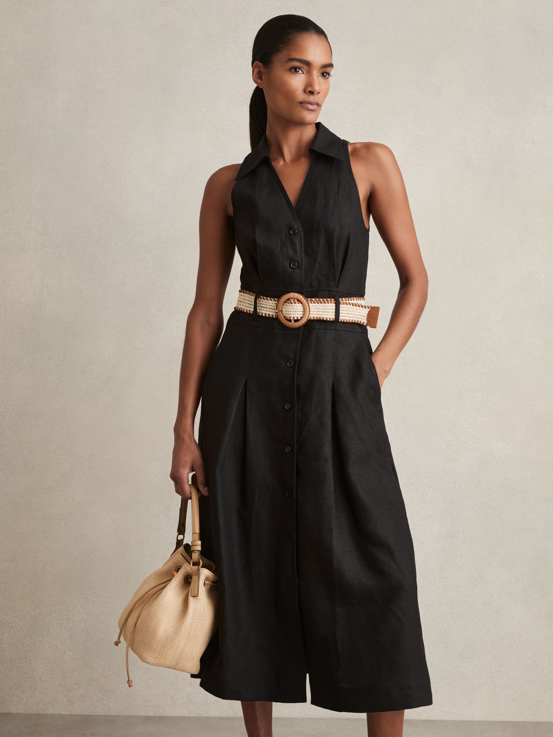 A woman wearing a black sleeveless linen midi dress with a V-neckline, collar, front buttons, pleated skirt, and a raffia woven belt with suede trim.