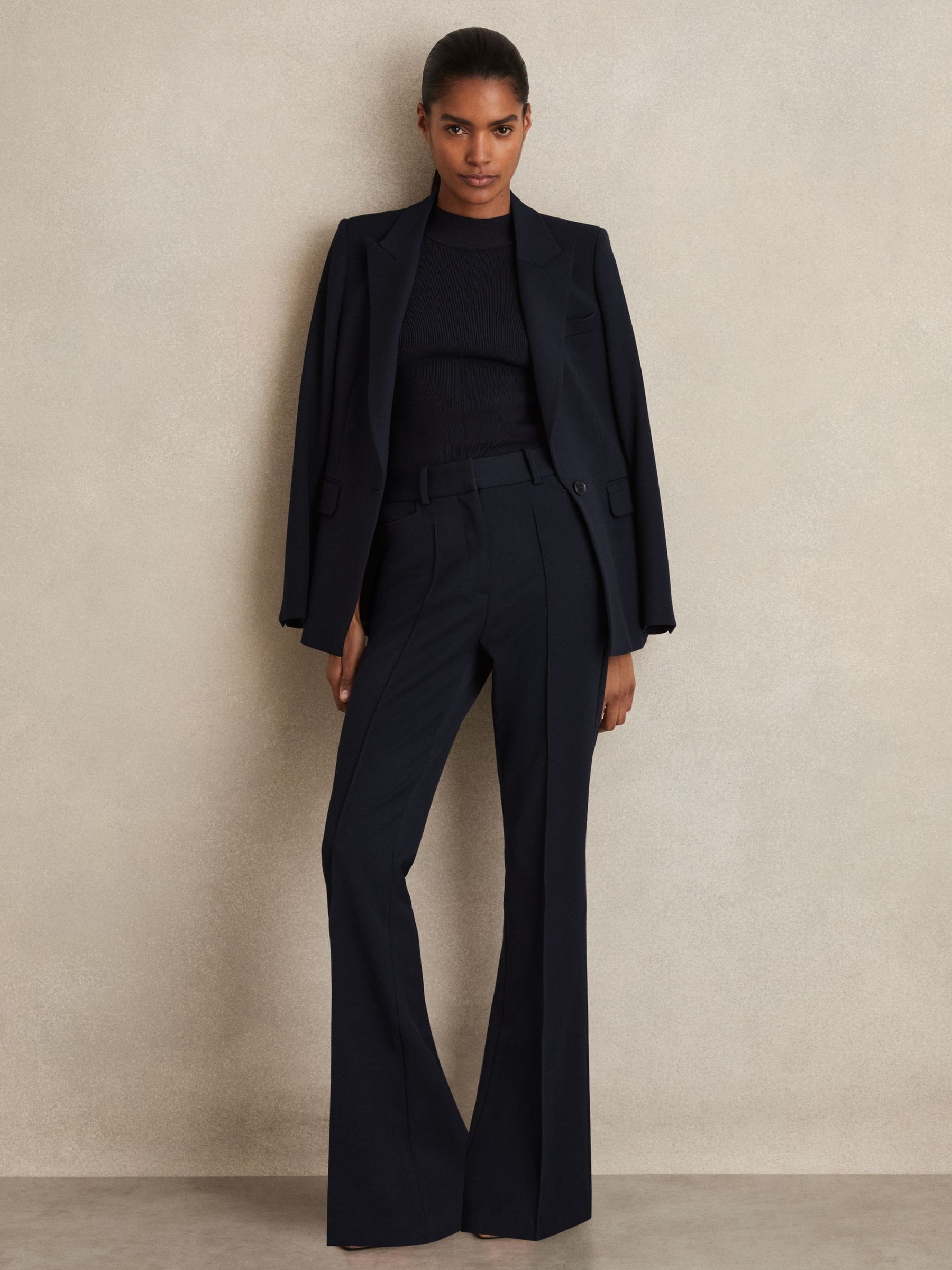 A woman wearing navy flared suit trousers with a black top and blazer, standing against a beige wall.