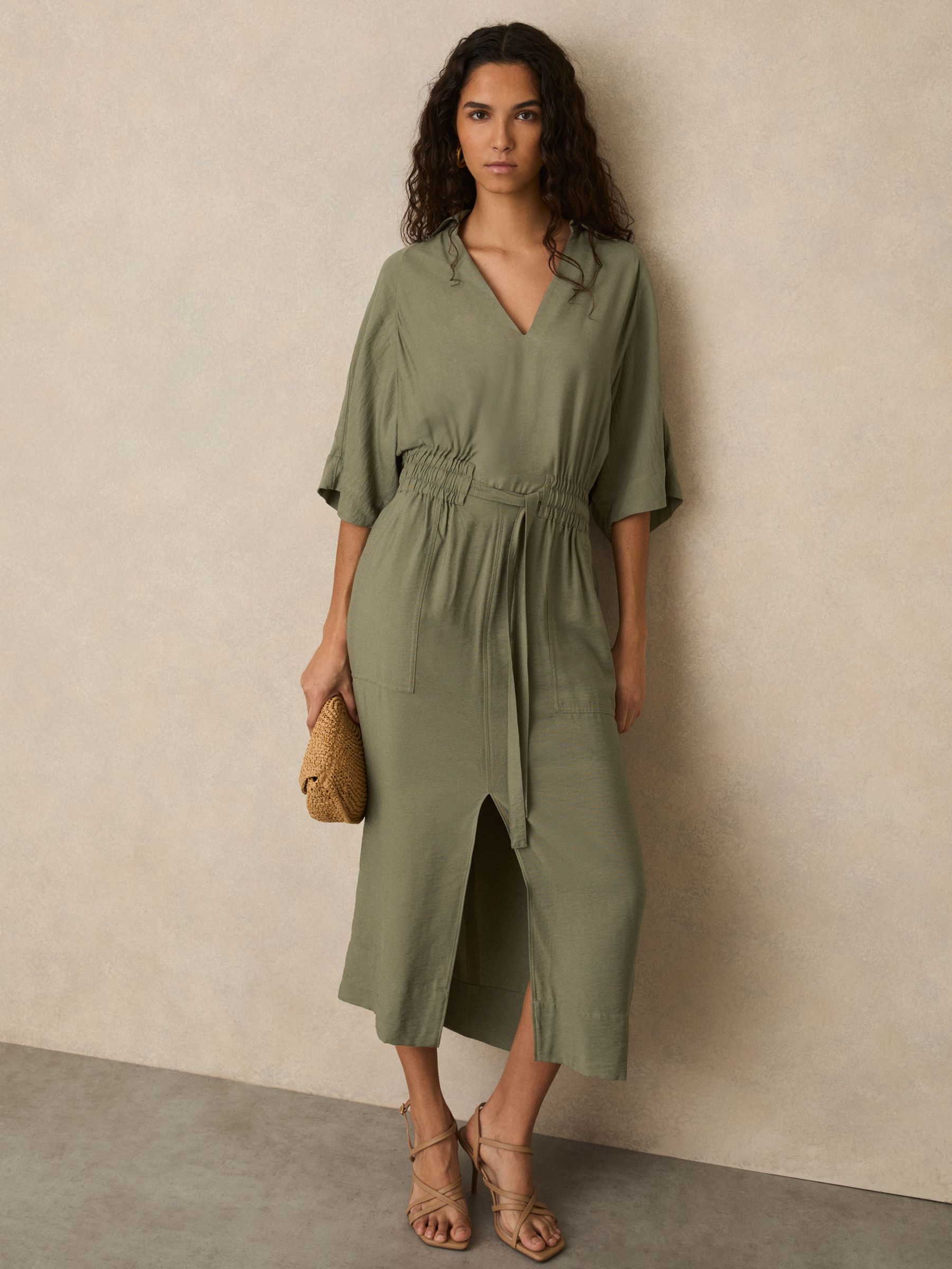A woman wearing a khaki midi shirt dress with a shirred waist, V-neckline, draped short sleeves, and a split front hem.