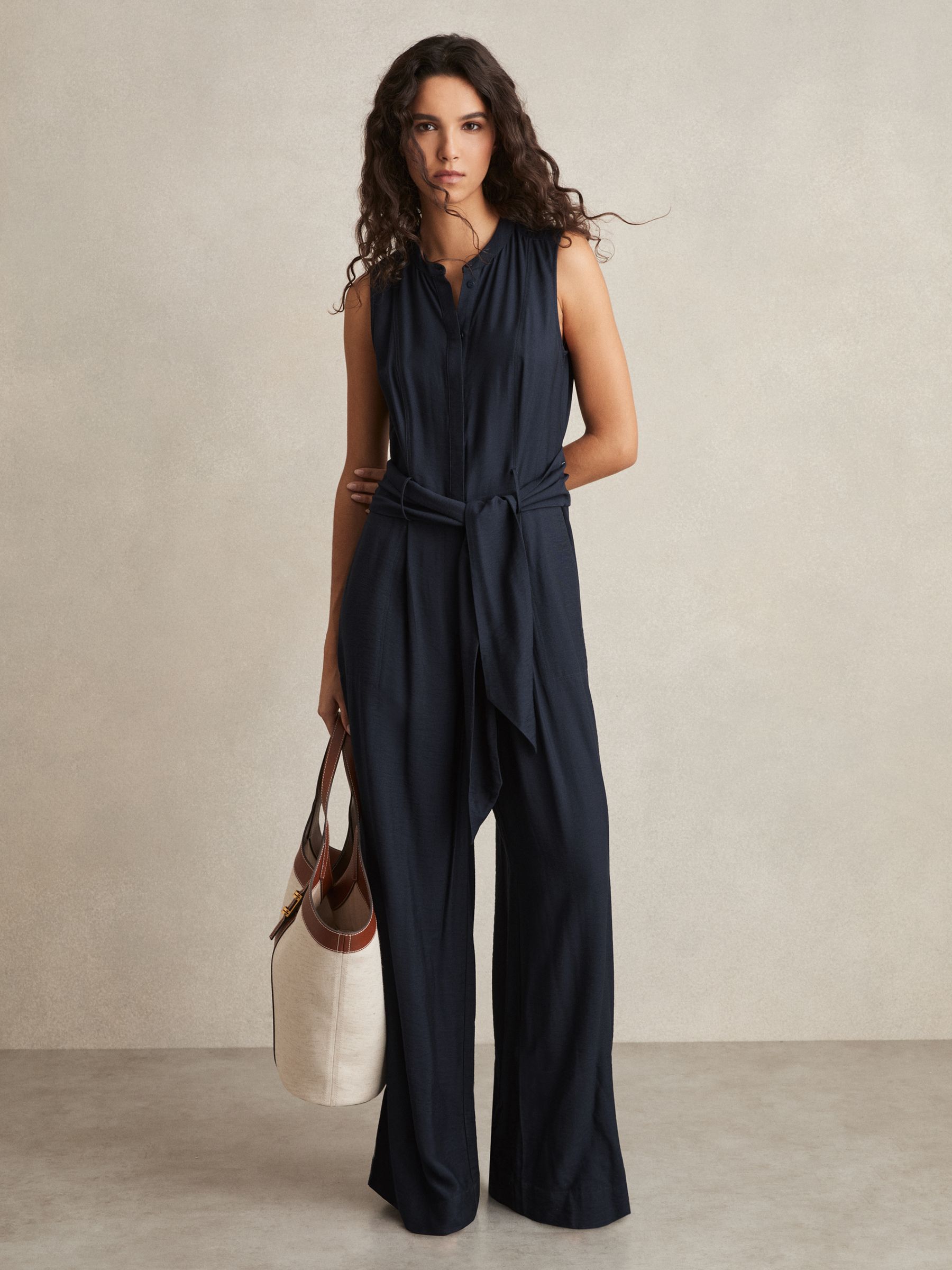 A woman wearing a navy wide-leg jumpsuit with a self-tie belt, band collar, and side pockets, holding a beige and brown tote bag.