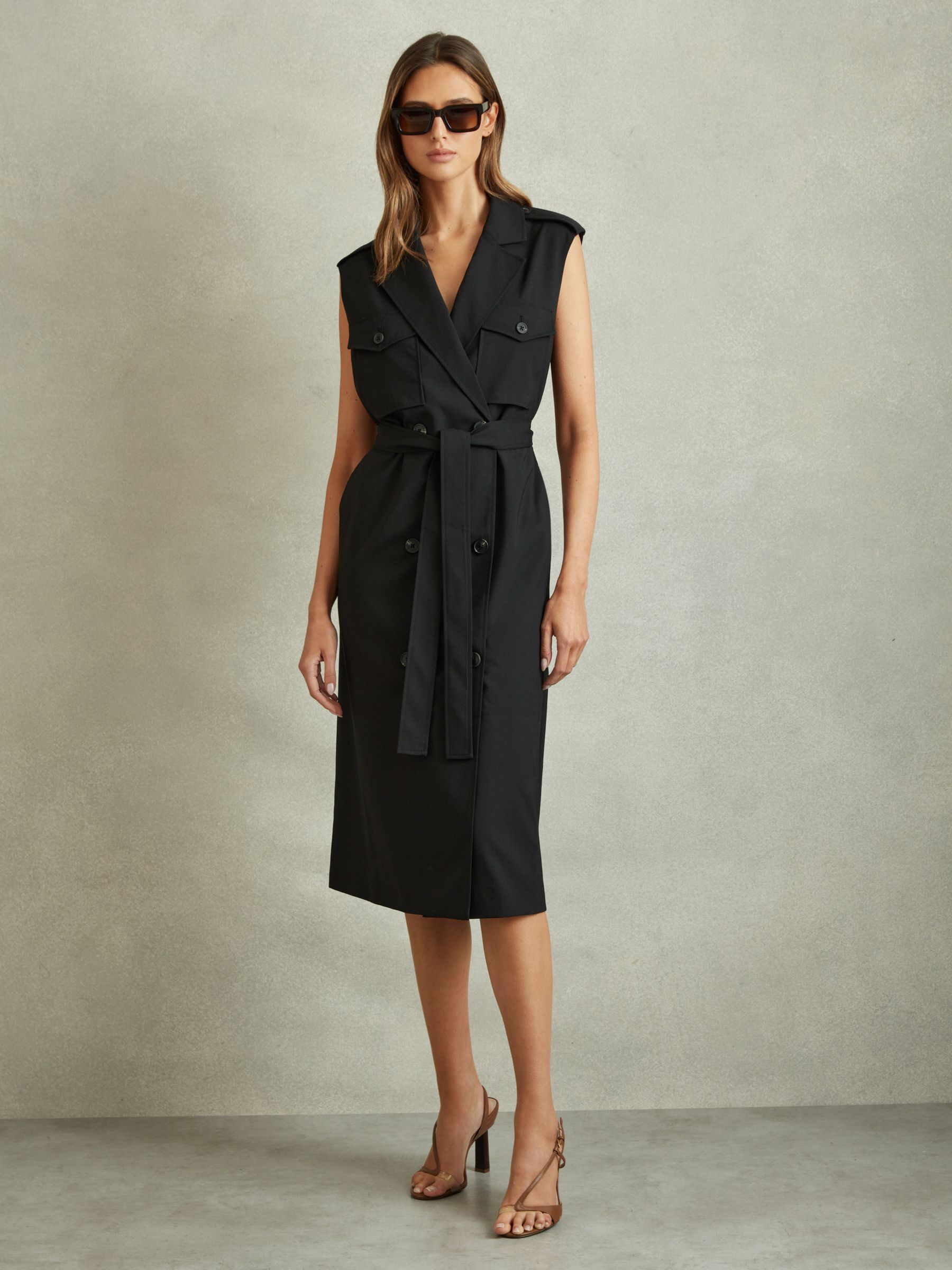 A woman wearing a black sleeveless wool-blend midi dress with double-breasted buttons, epaulettes, and a waist belt, standing against a plain background.