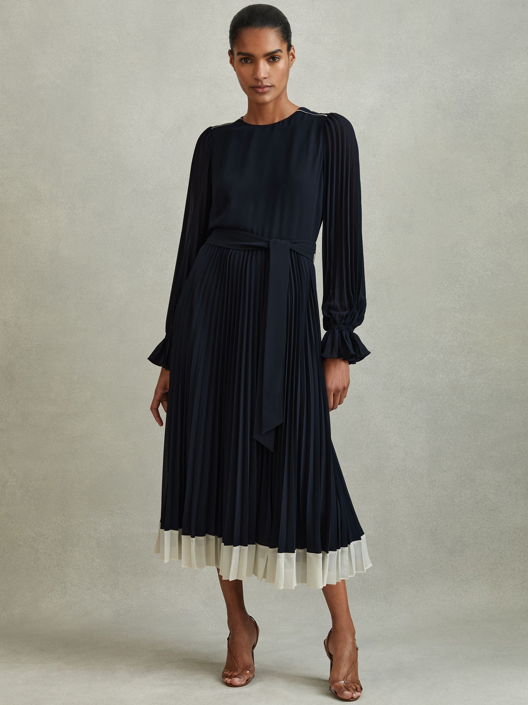 A woman wearing a navy and cream pleated midi dress with sheer long sleeves, elasticated cuffs, a self-tie waist belt, and a zip at the back.