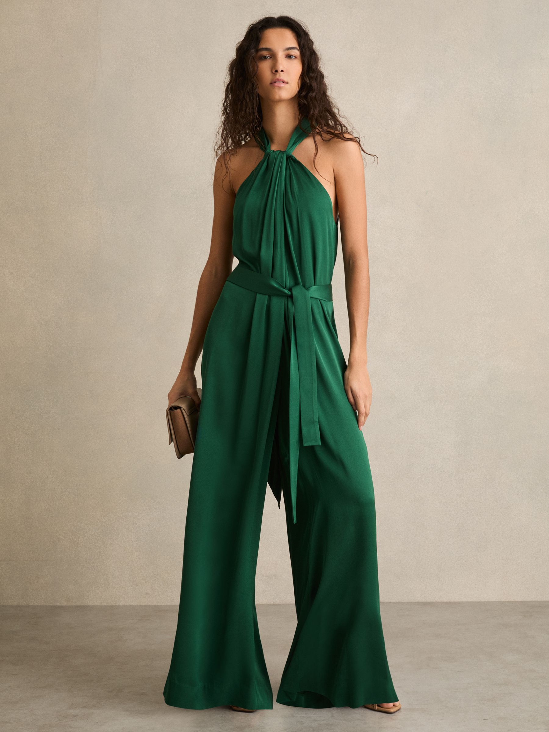 A woman wearing a green satin wide-leg draped jumpsuit with a halter neckline, self-tie sash at the back, and side pockets.