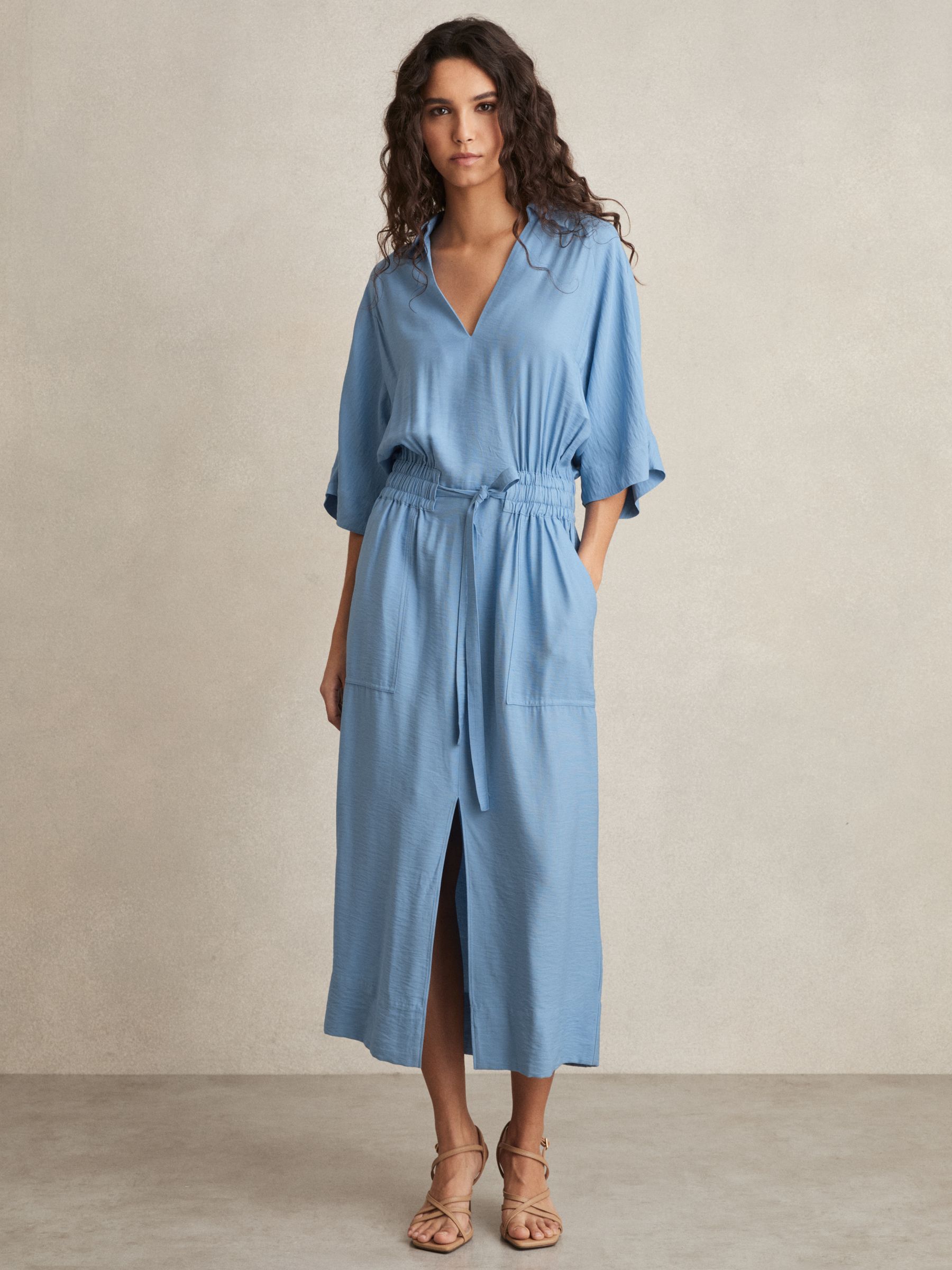 A woman wearing a blue midi shirt dress with a shirred waist, V-neckline, and draped sleeves.