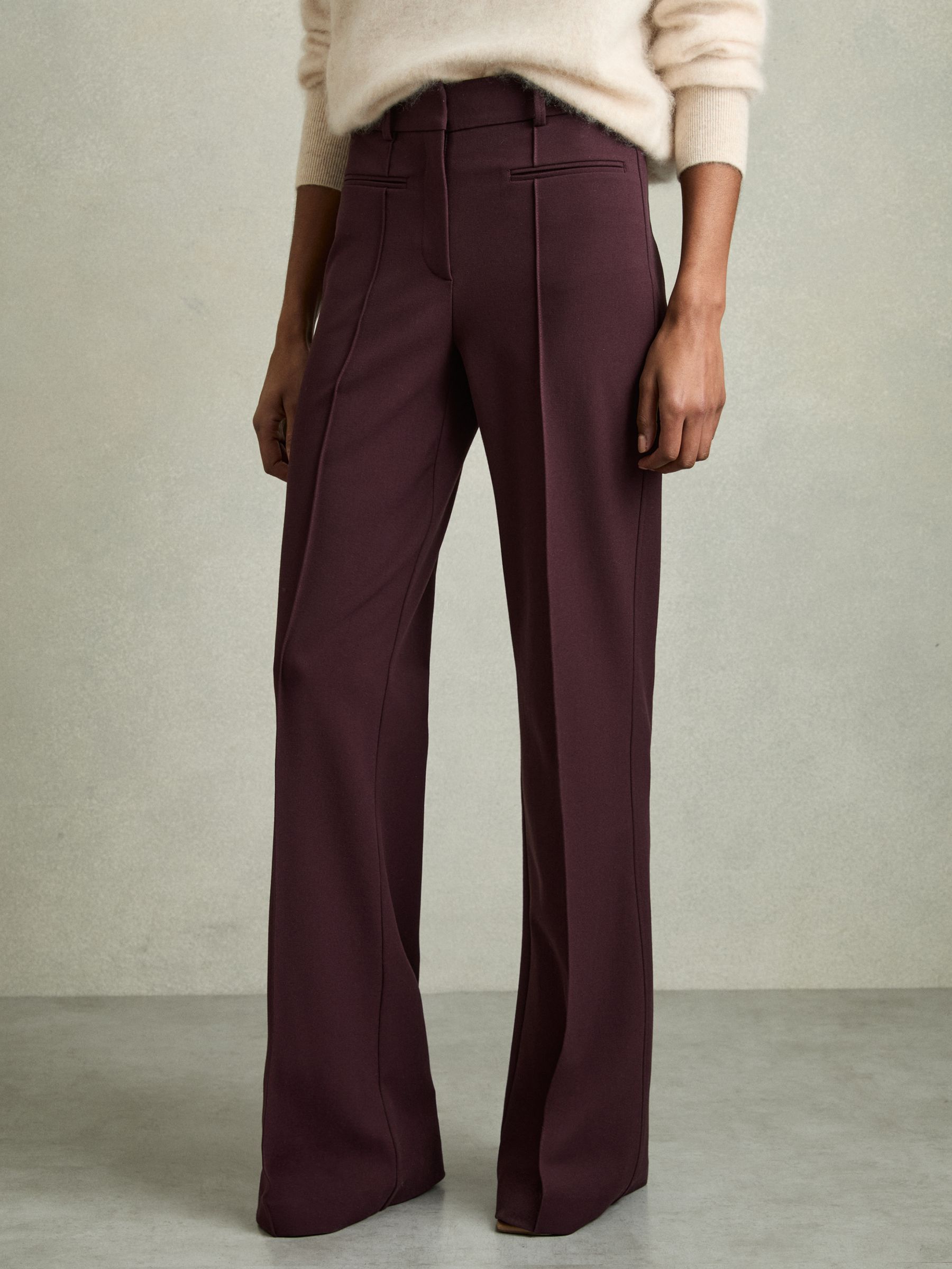 A pair of burgundy women's trousers with a high waist, pinched front seams, and a flared leg, shown on a model.