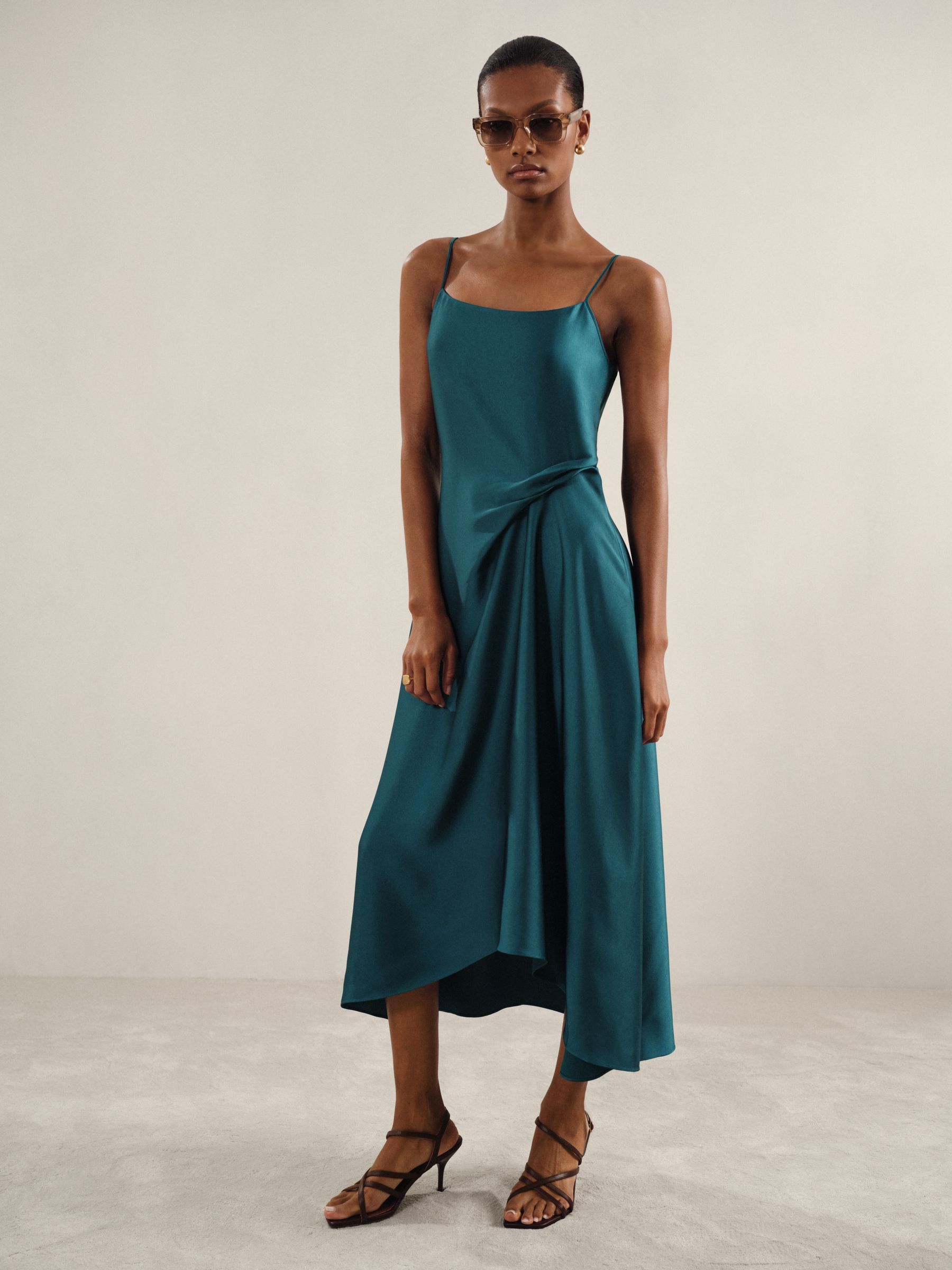 A woman wearing a teal satin slip dress with drape detail, adjustable shoulder straps, and a midi-length asymmetric hem.