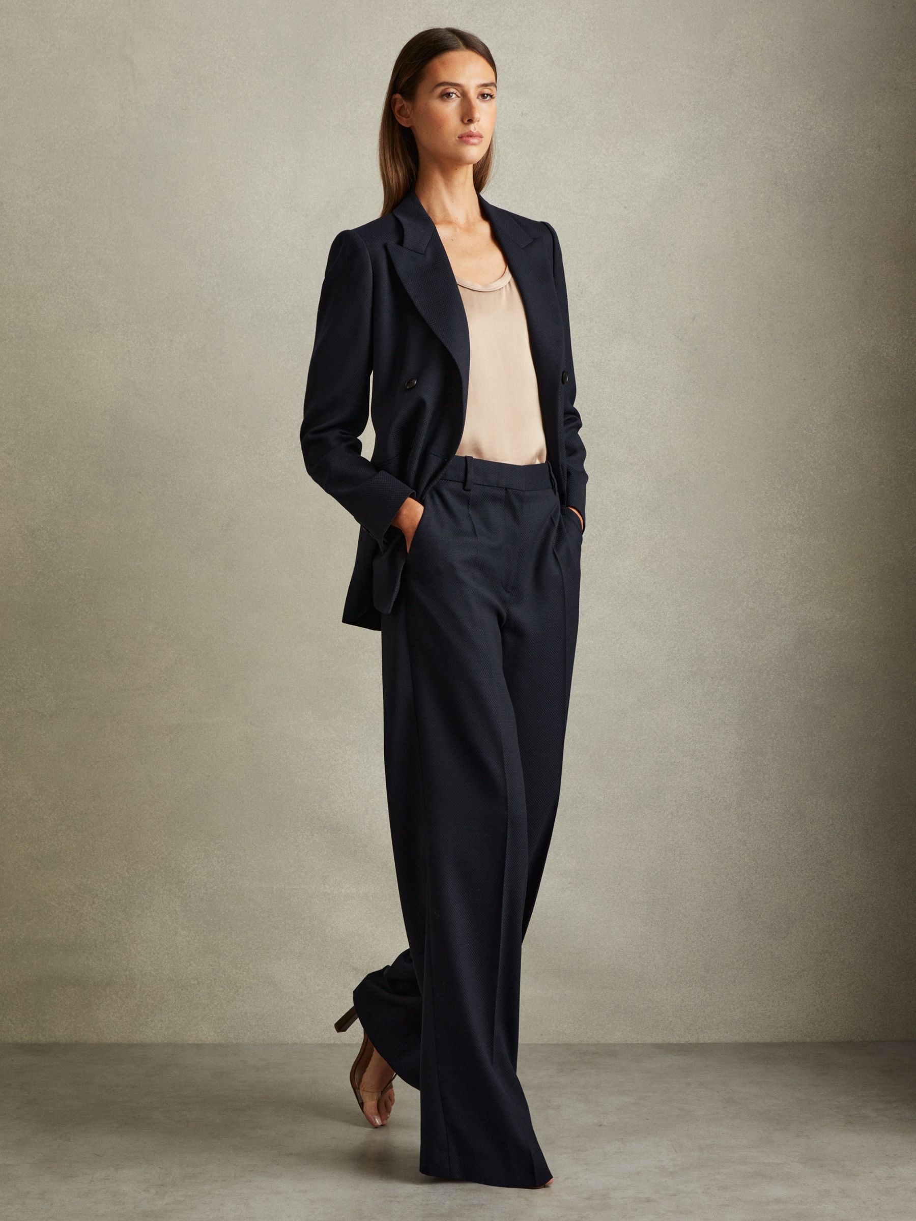 A woman wearing navy textured wide-leg trousers with a matching blazer and beige top, standing with her hands in her pockets.