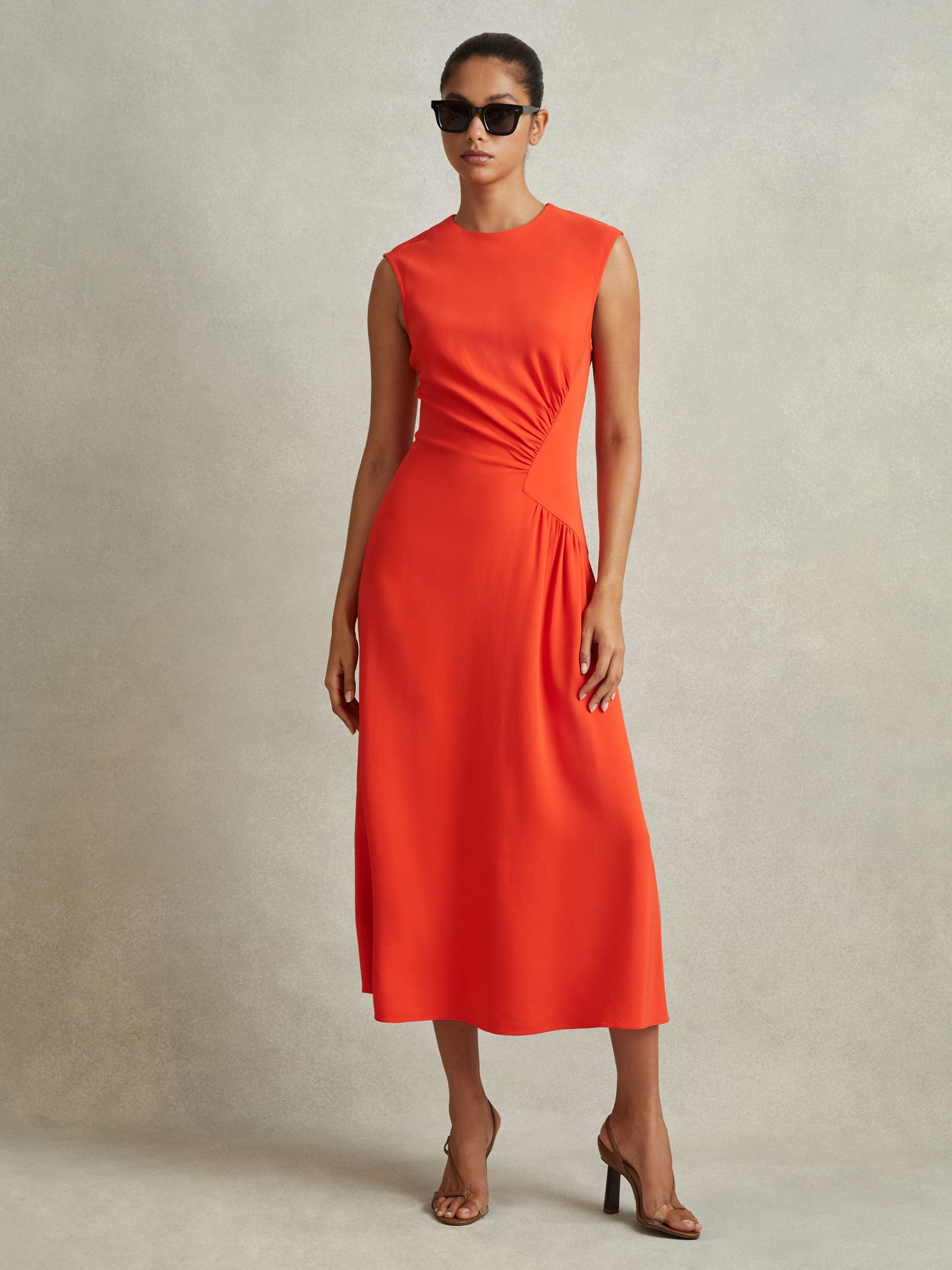 A woman wearing a sleeveless, orange midi dress with ruched detailing at the waist, a round neckline, and a zip closure at the back. She is also wearing sunglasses and high heels.