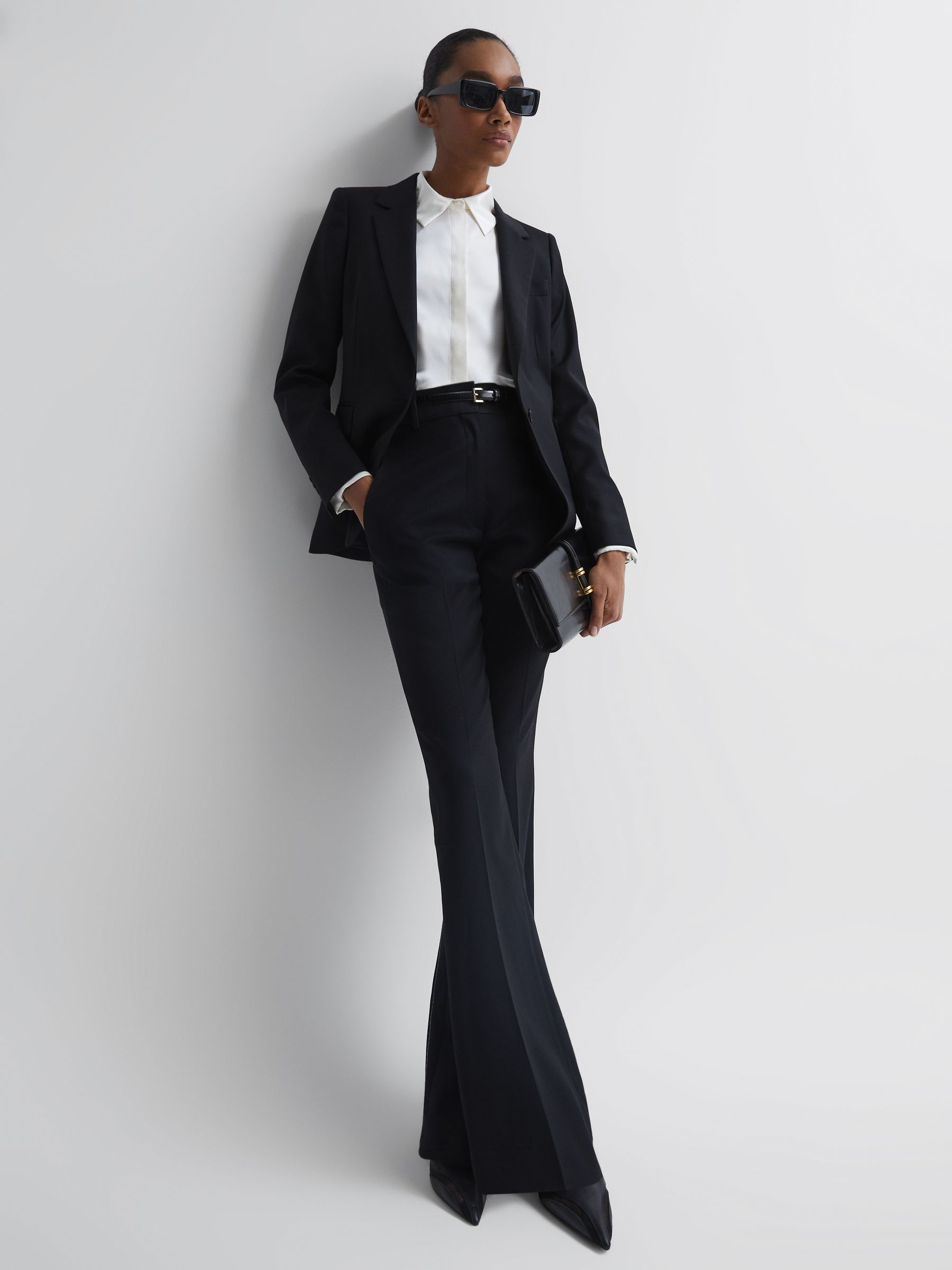 A woman wearing black tailored flared trousers with a white shirt and black blazer, holding a black clutch, and wearing sunglasses.