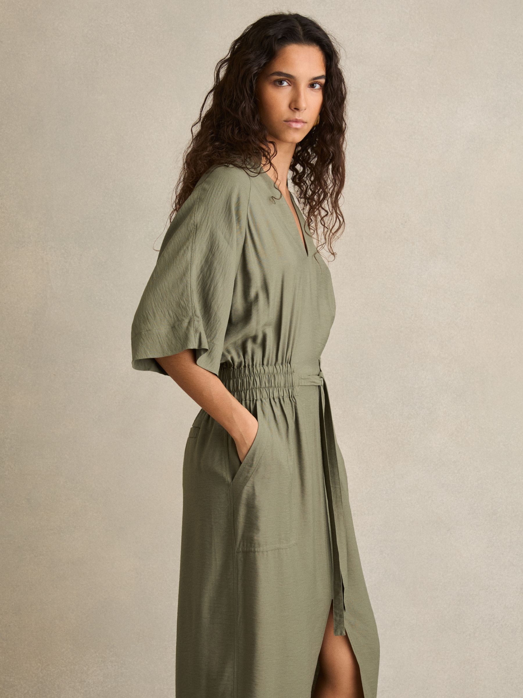 A woman wearing a khaki midi shirt dress with a shirred waist, V-neckline, and draped short sleeves, standing against a plain background.