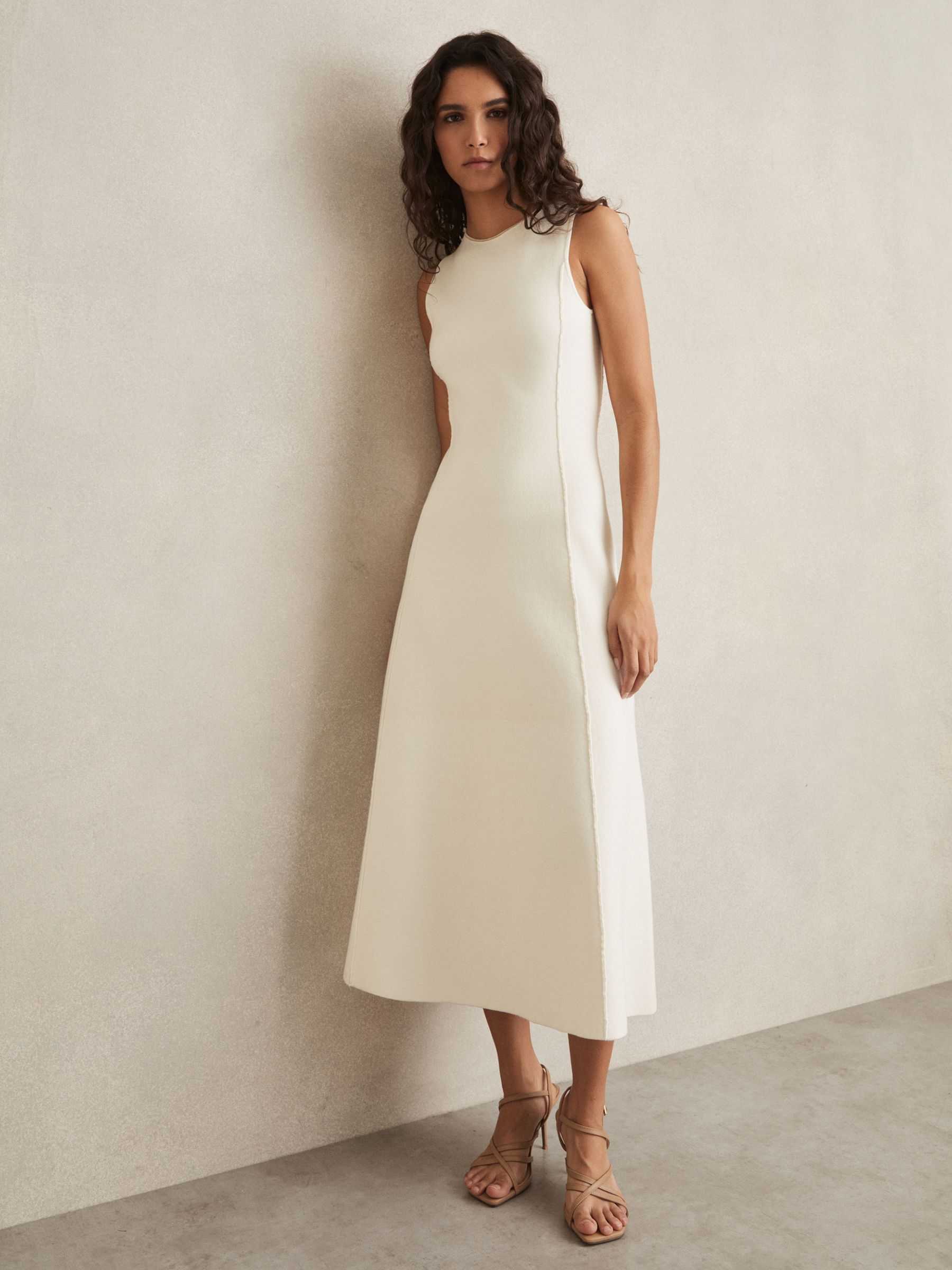 A woman wearing an ivory knitted midi dress with seam details and a metallic embroidered neckline, standing against a plain wall.