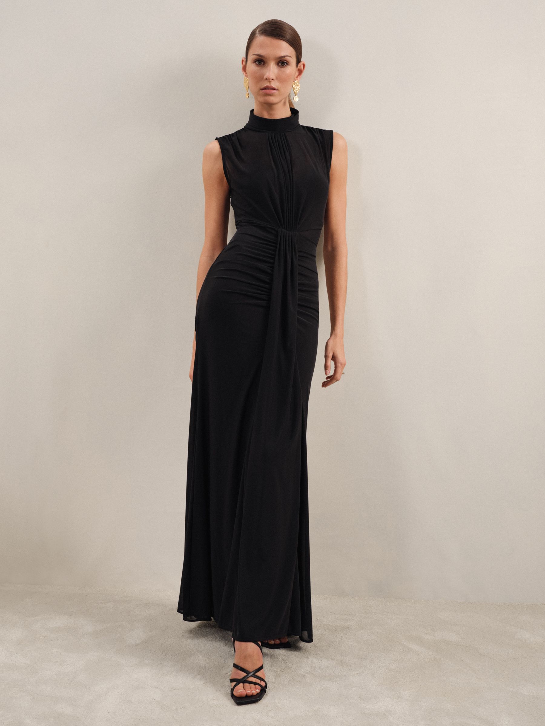 A woman wearing a black sheer ruched maxi dress with a high neckline and sleeveless design.