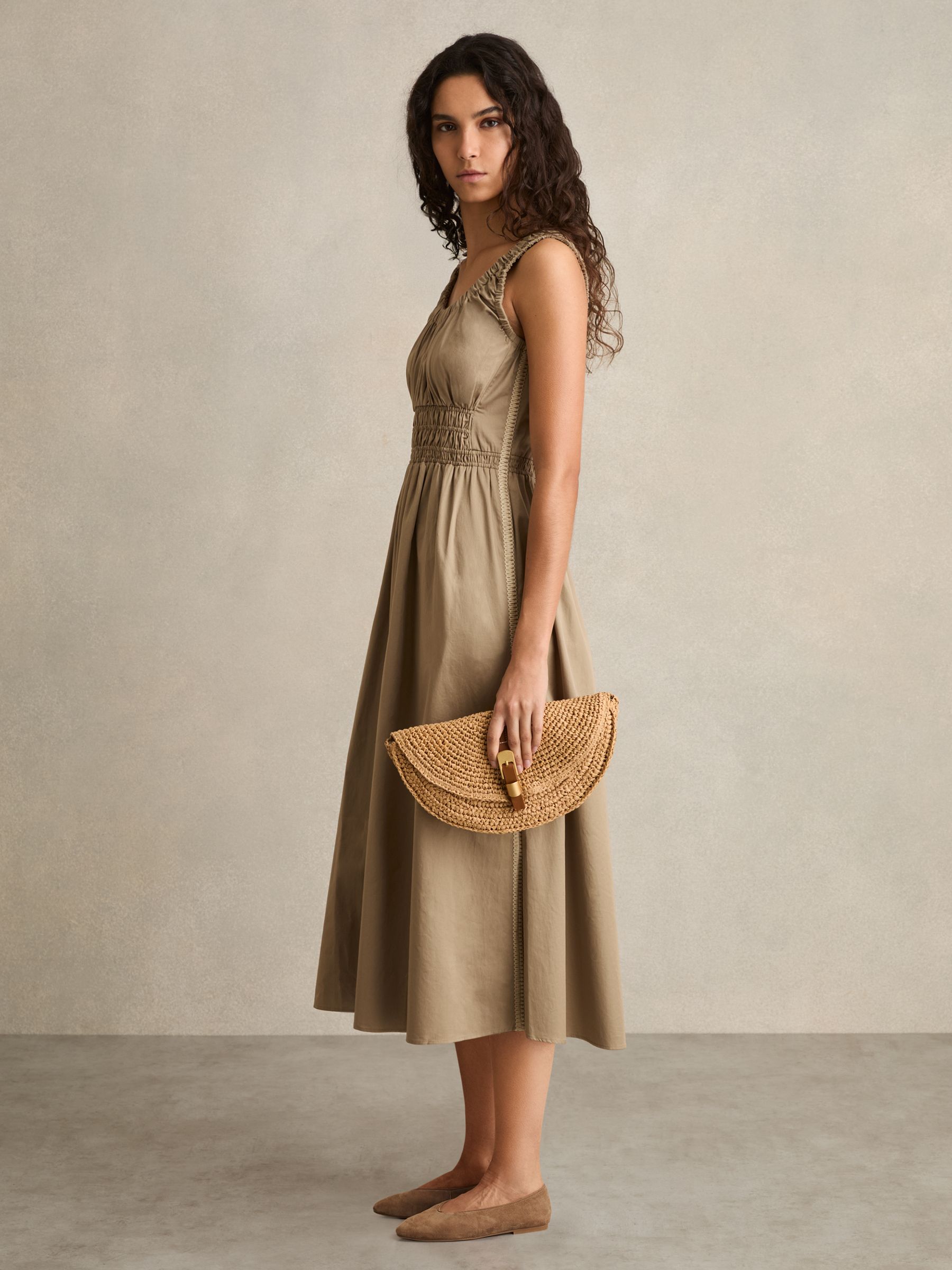 A woman wearing a khaki midi dress with shirring at the waist, craft stitching on the sides, a scoop neckline, and side slits at the hem.