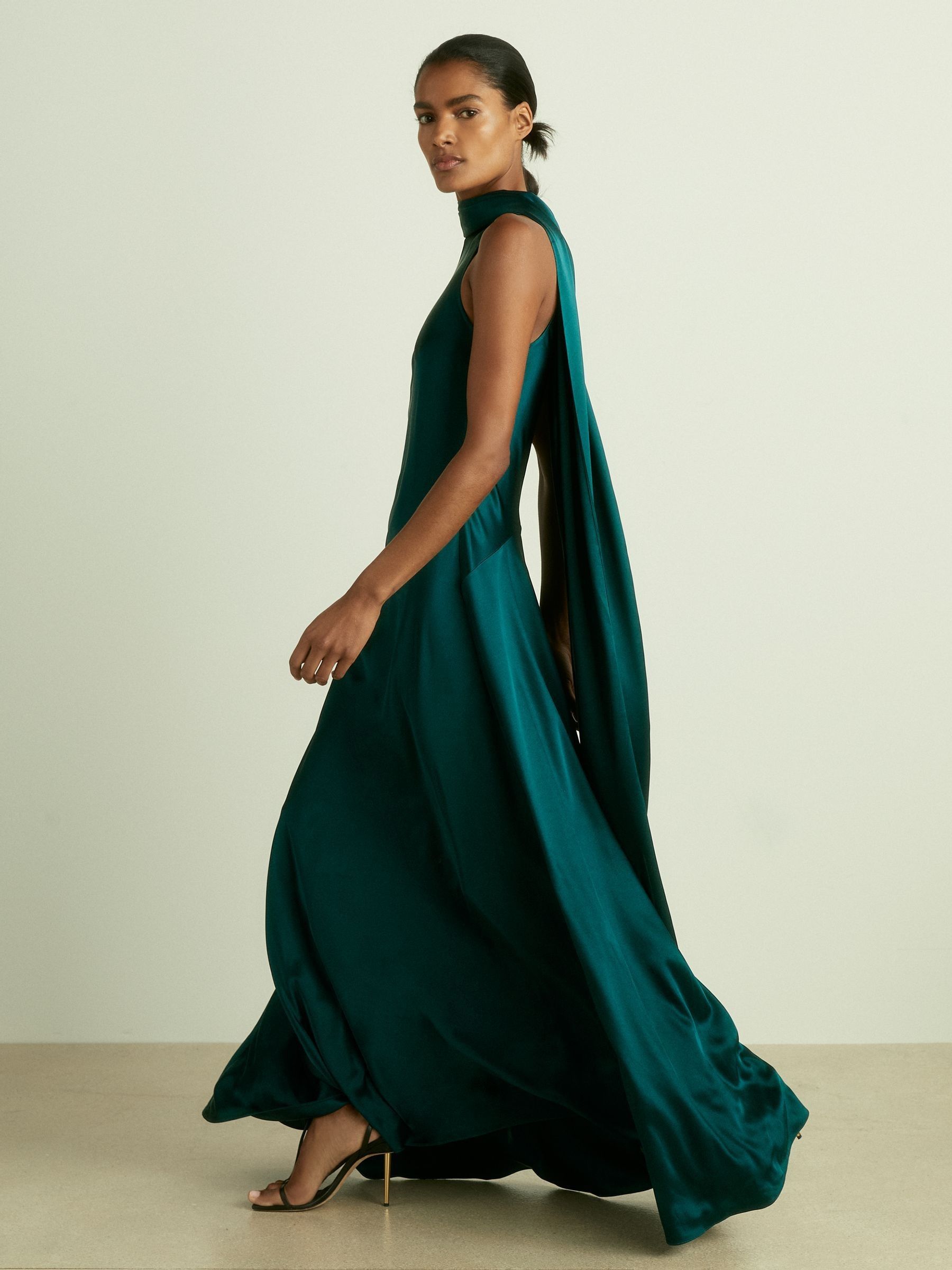A woman wearing a teal satin maxi dress with a high neck and a cape detail draping down the back, standing against a plain background.