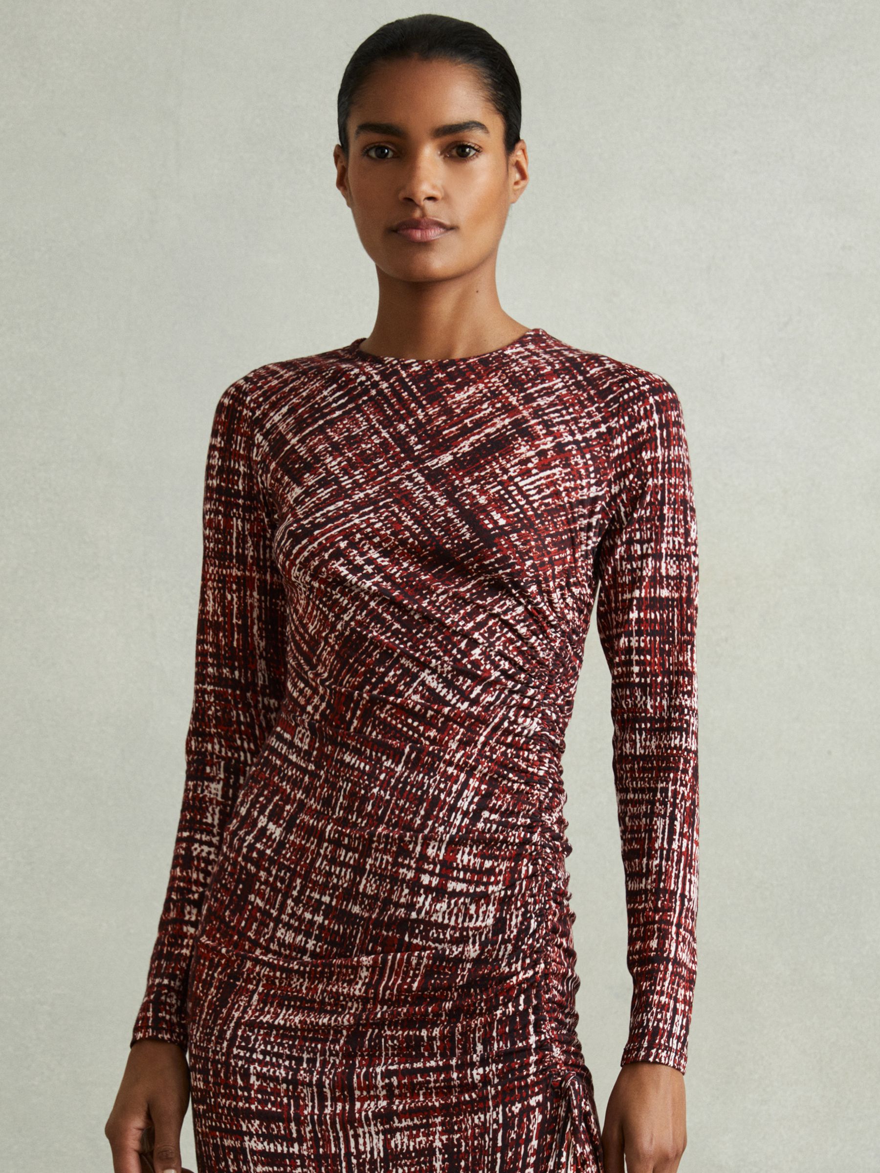 A woman wearing a red midi dress with a speckled print, ruched side seam with a drawstring, long sleeves, and a side split.