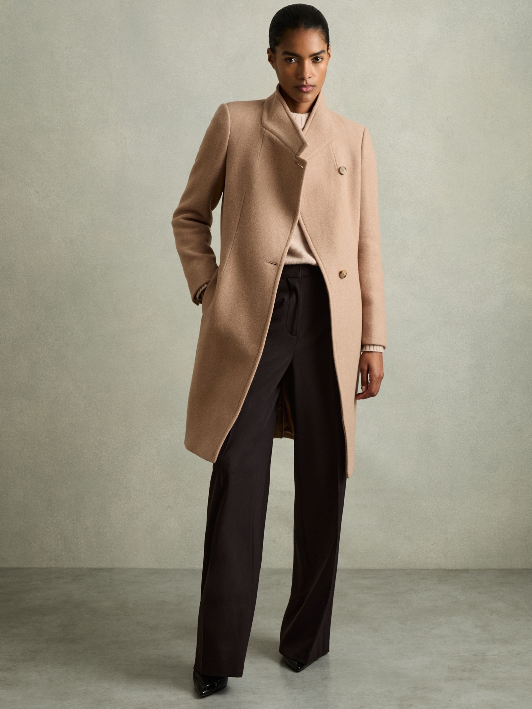 A woman wearing a light camel wool blend longline double-breasted coat with notched lapels, styled with black trousers and a beige top.