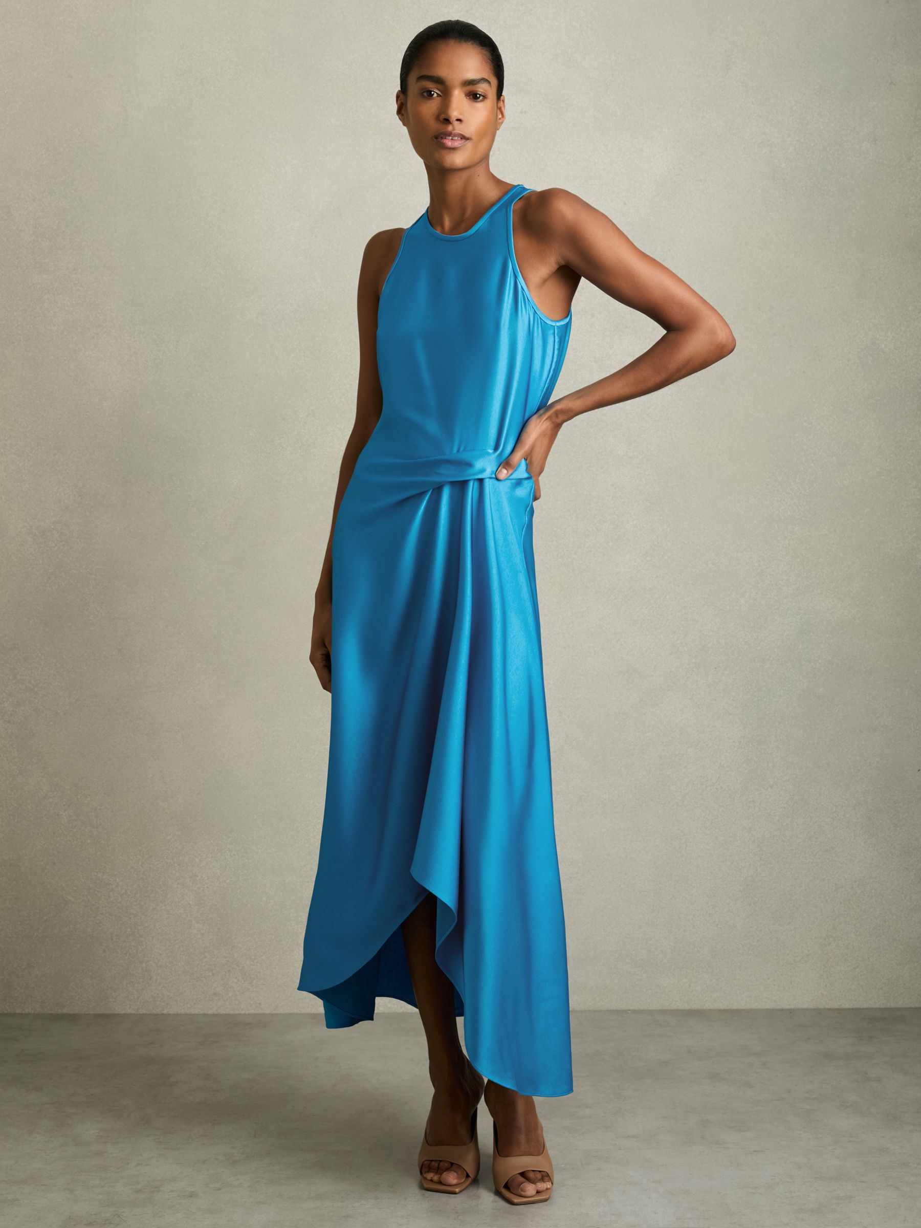 A woman wearing a blue satin midi dress with a draped design, tuck detail at the waist, asymmetric hem, and keyhole back.