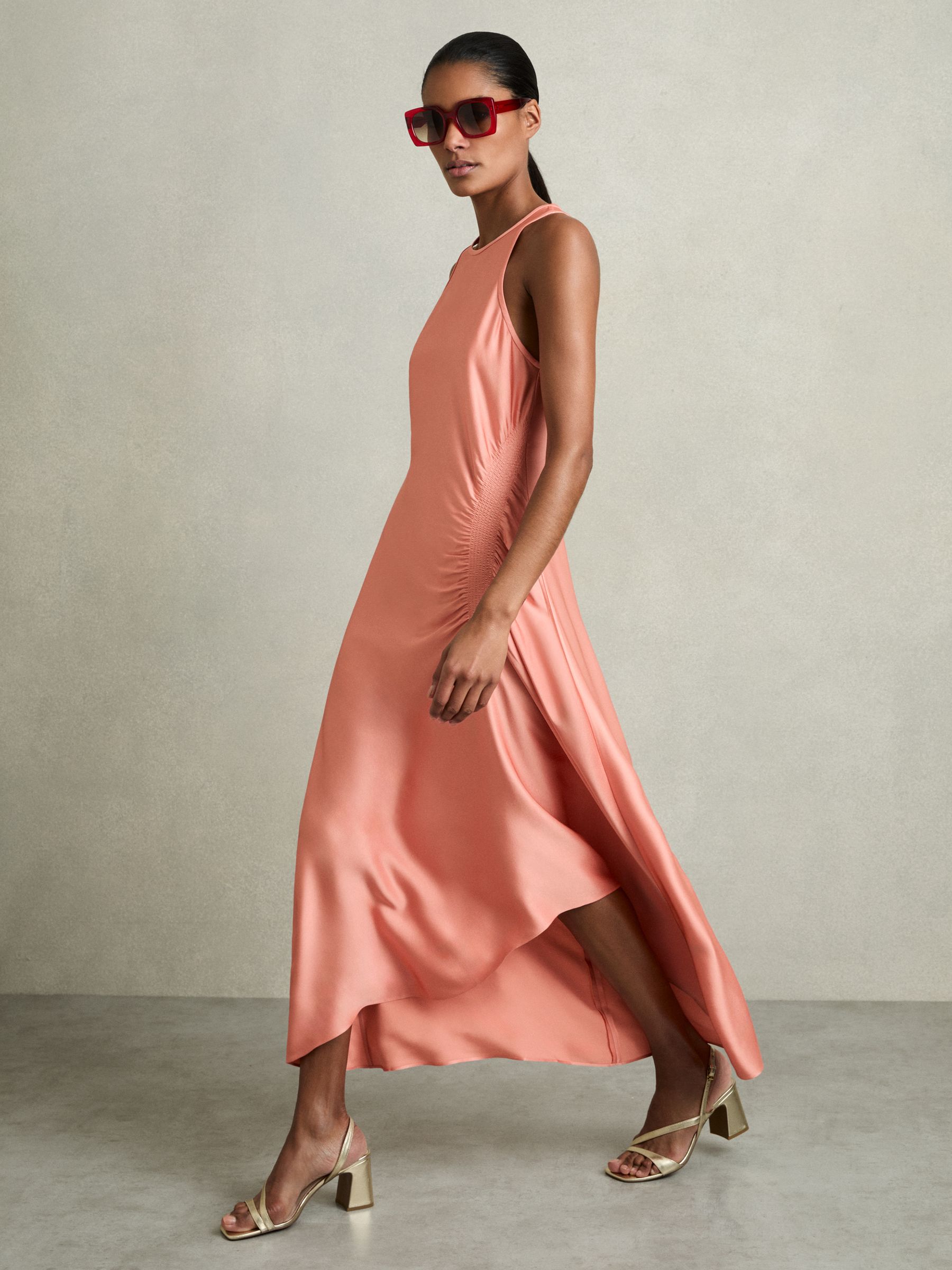 A woman wearing a coral satin midi dress with shirred sides, high-low hem, and a round neckline, paired with beige high-heeled sandals and red sunglasses.
