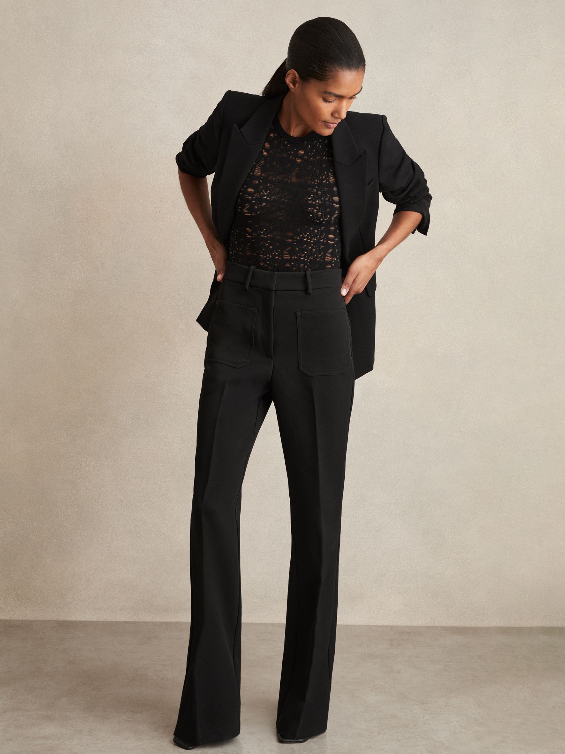 A woman wearing black high-rise flared trousers with patch pockets, a hook-and-bar fastening, and a zip, paired with a black blazer and a black lace top.