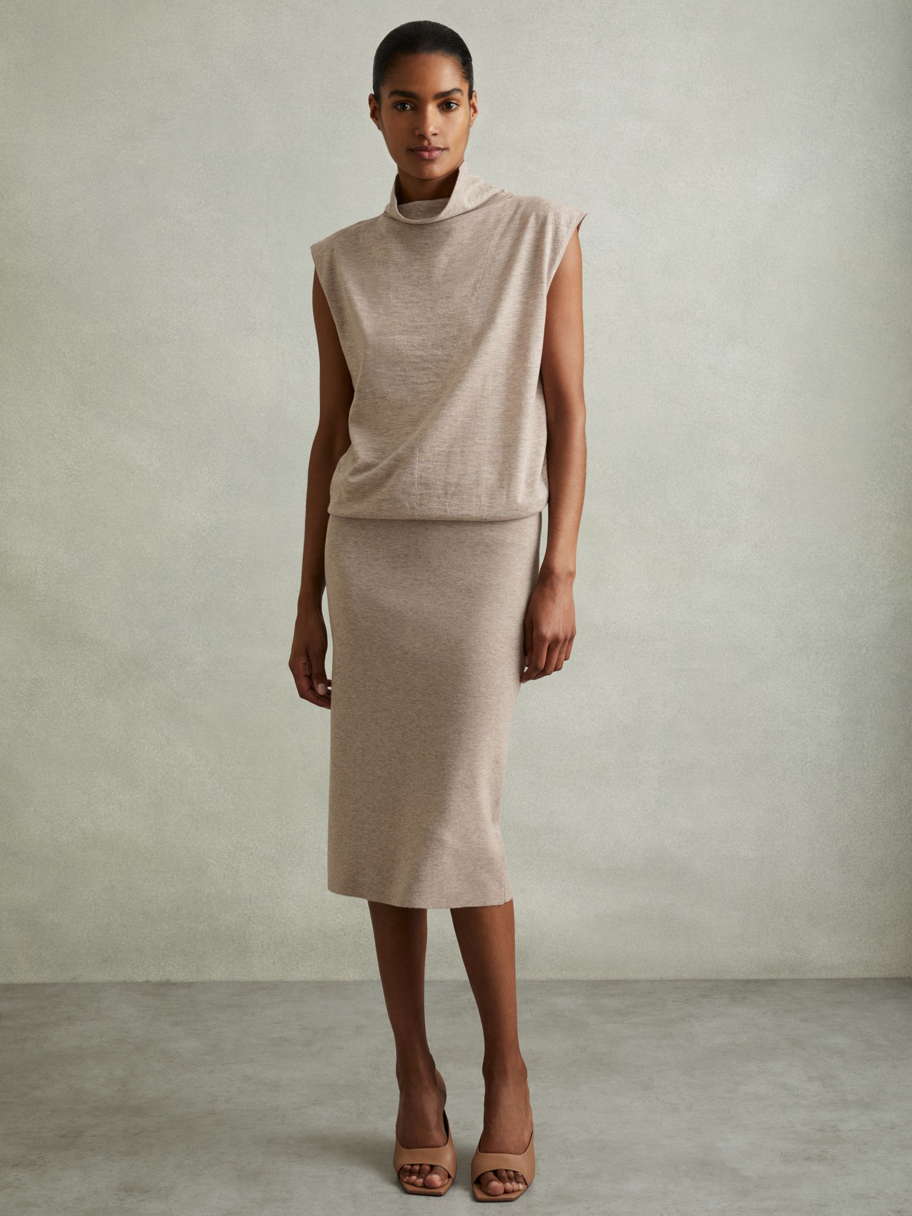 A woman wearing a beige knitted wool-cashmere midi dress with a funnel neckline, blouson top, cap sleeves, and a fitted skirt.