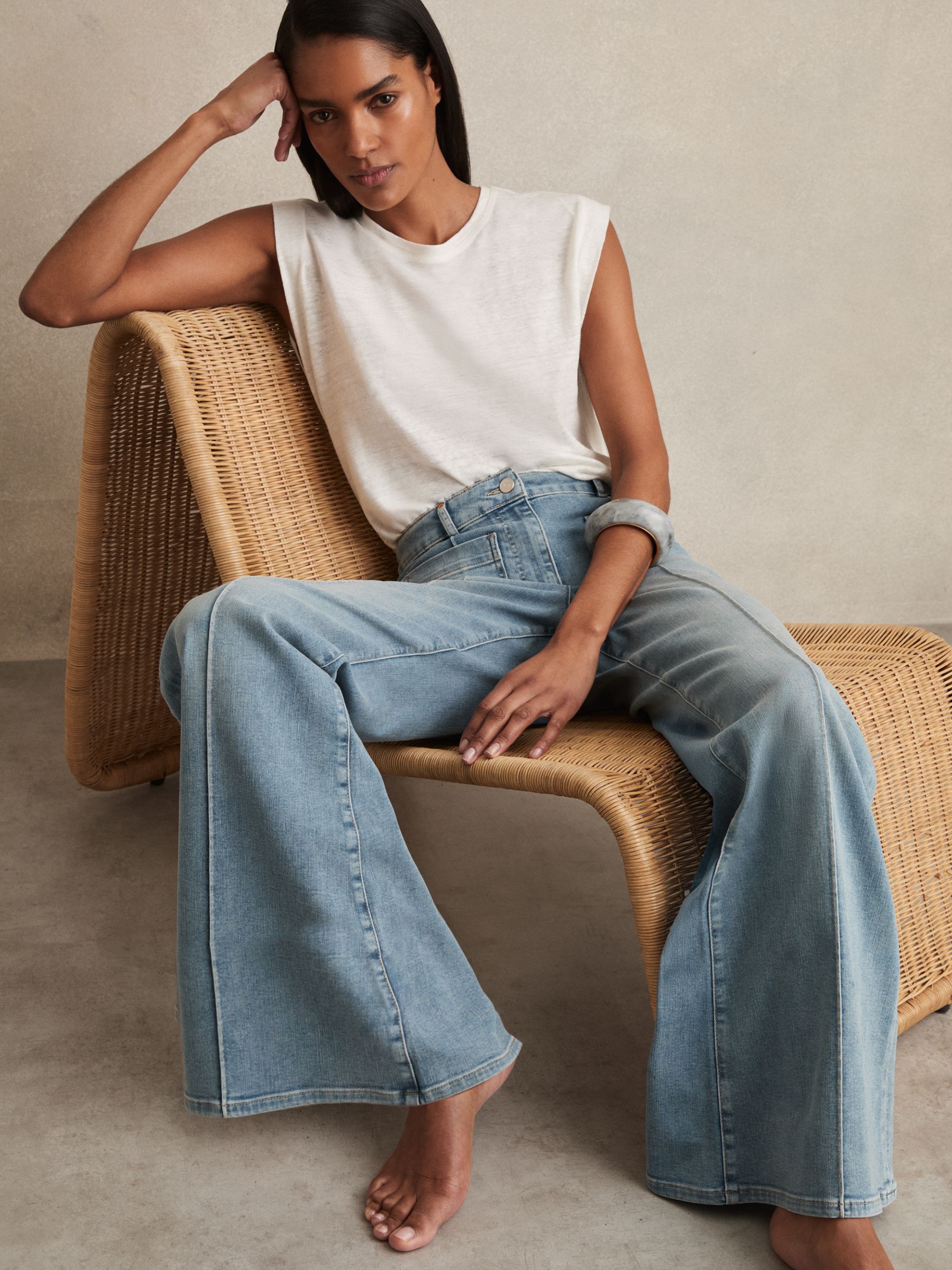 A woman wearing light blue flared jeans with patch pockets and a white sleeveless top, sitting on a wicker chair.