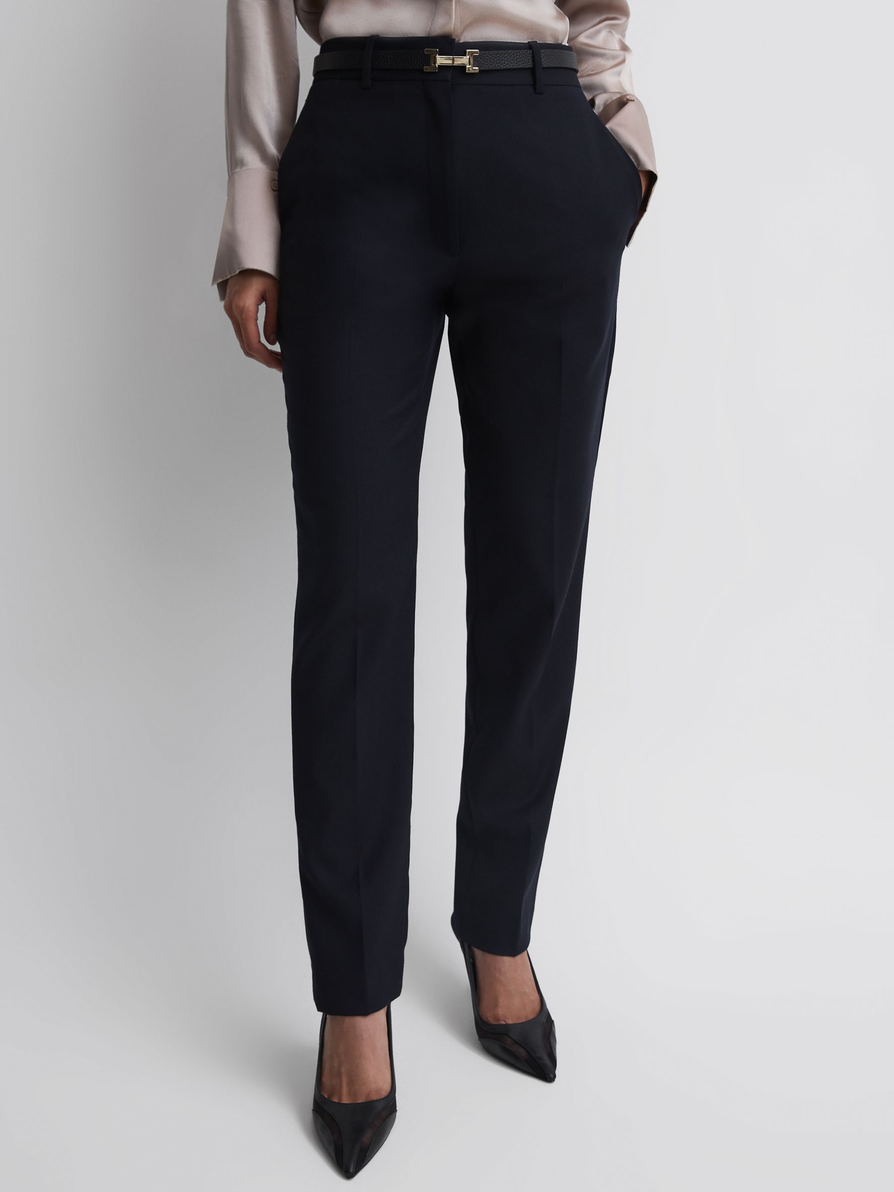 A pair of navy women's tapered suit trousers with side pockets and pressed creases, styled with a beige blouse and black heels.