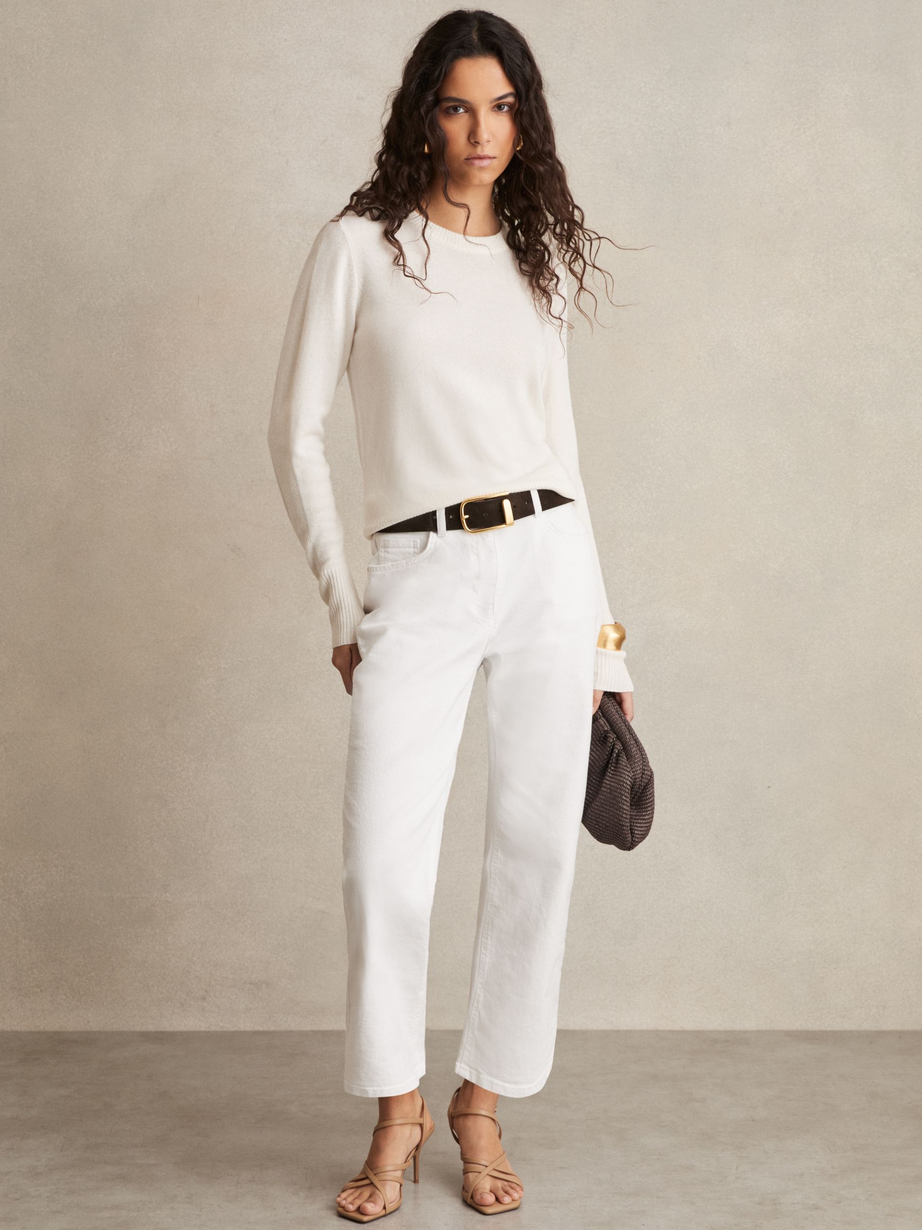 A woman wearing white petite mid-rise straight-leg jeans, a white top, and beige high-heeled sandals, holding a small brown bag.