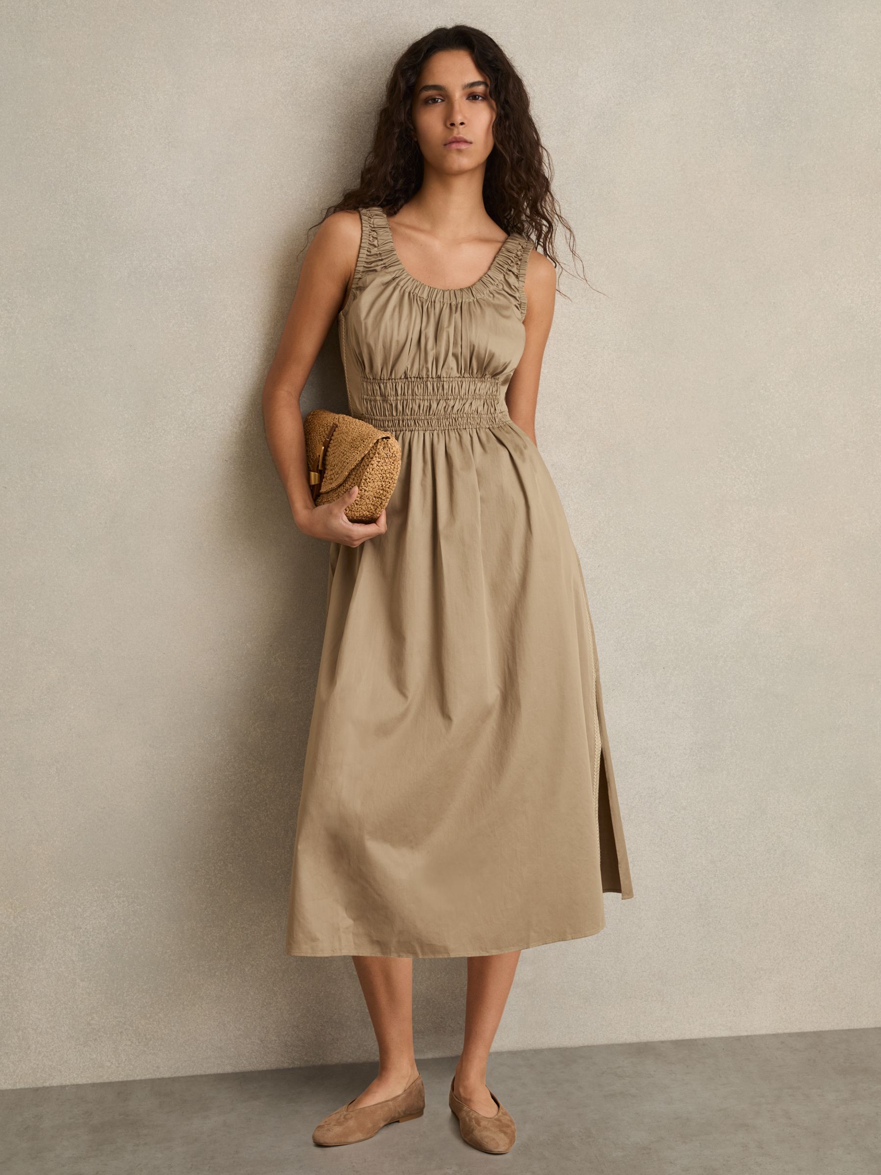A woman wearing a khaki midi dress with a shirred waist, craft stitching on the sides, a scoop neckline, and side slits at the hem.