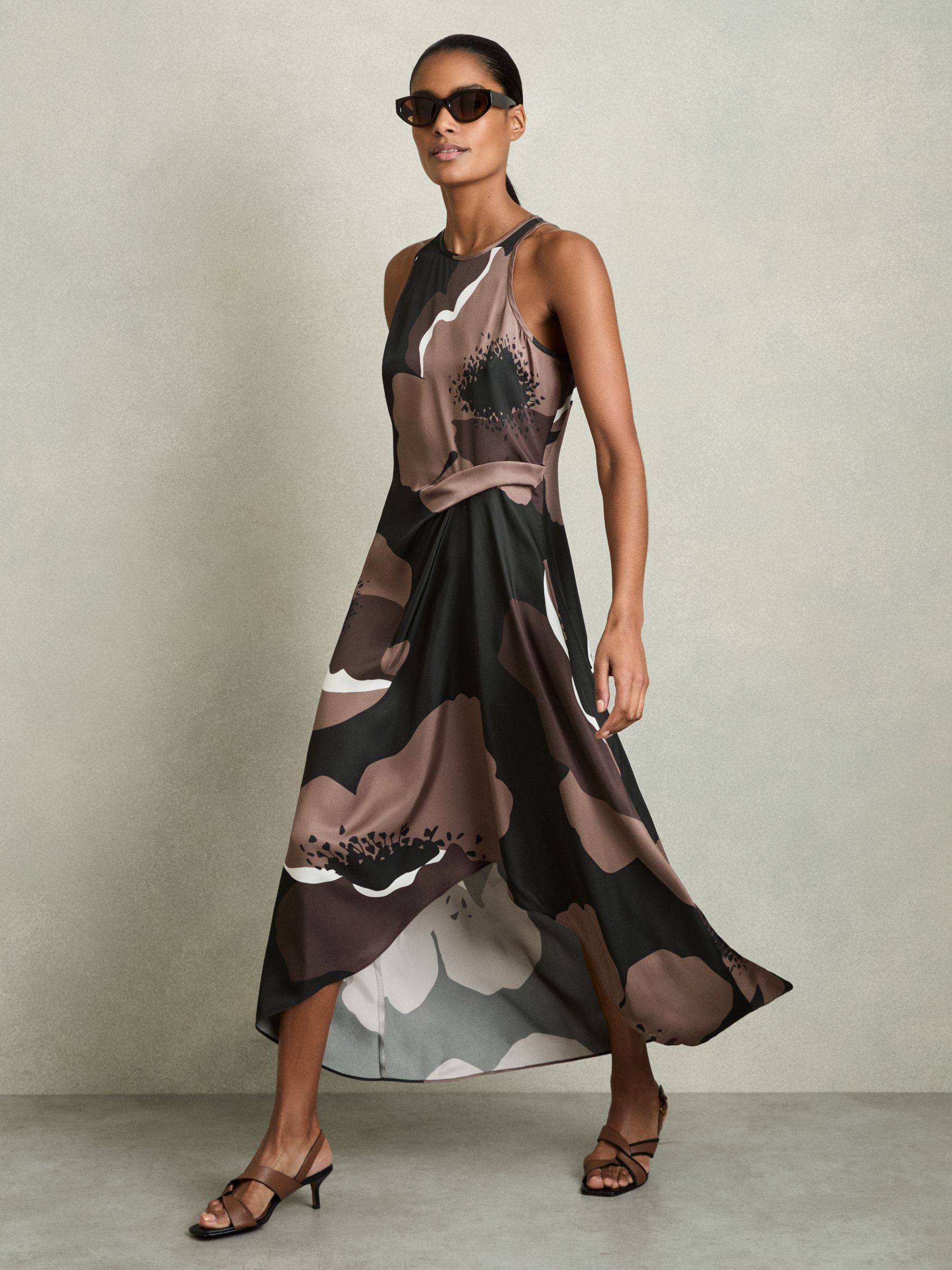 A woman wearing a satin floral midi dress in brown with a keyhole back and asymmetric hem, paired with sunglasses and heels.