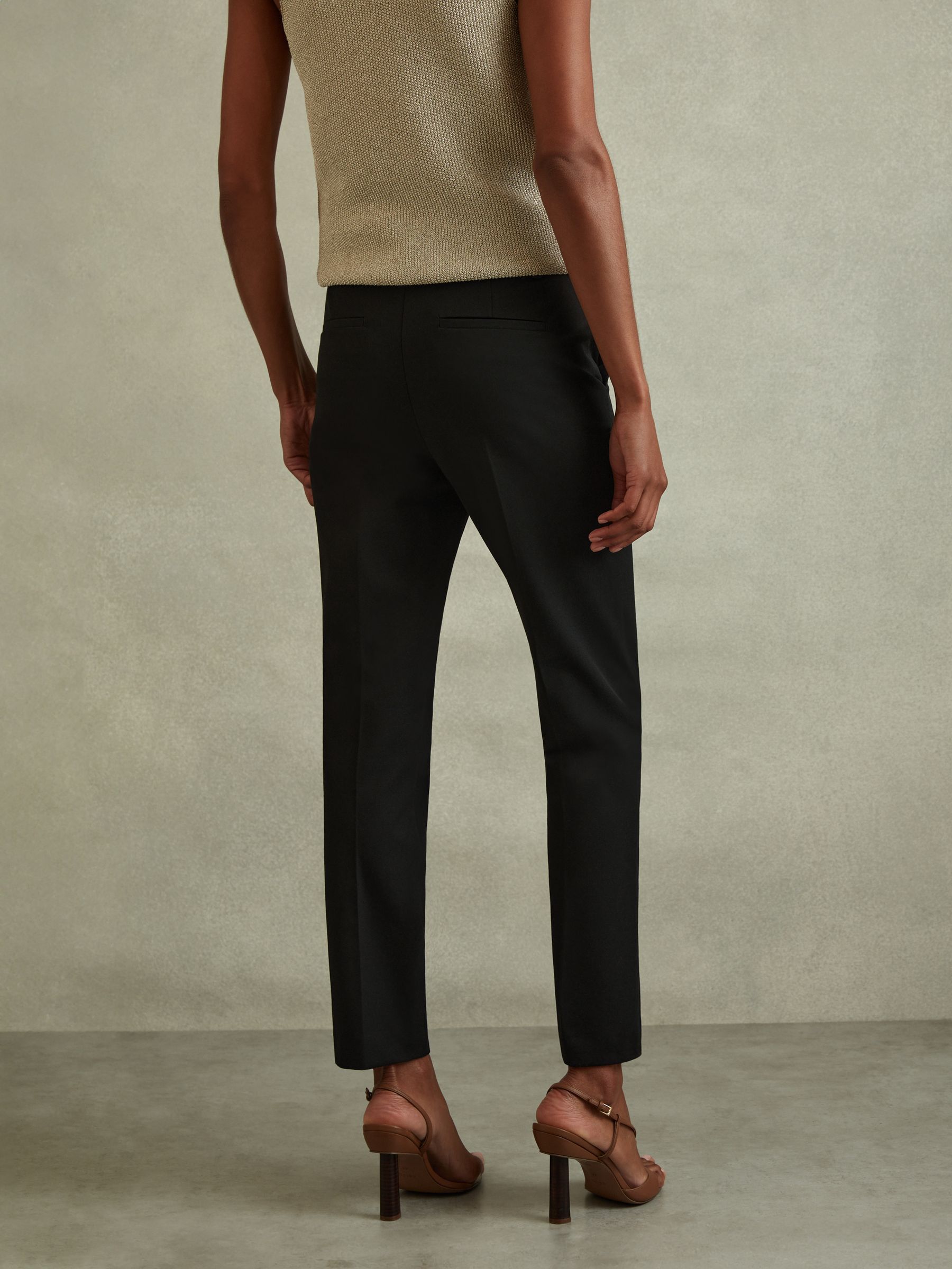 A woman wearing black tailored slim fit trousers with side and back pockets, a hook and zip fastening, and ankle length, paired with beige heels.