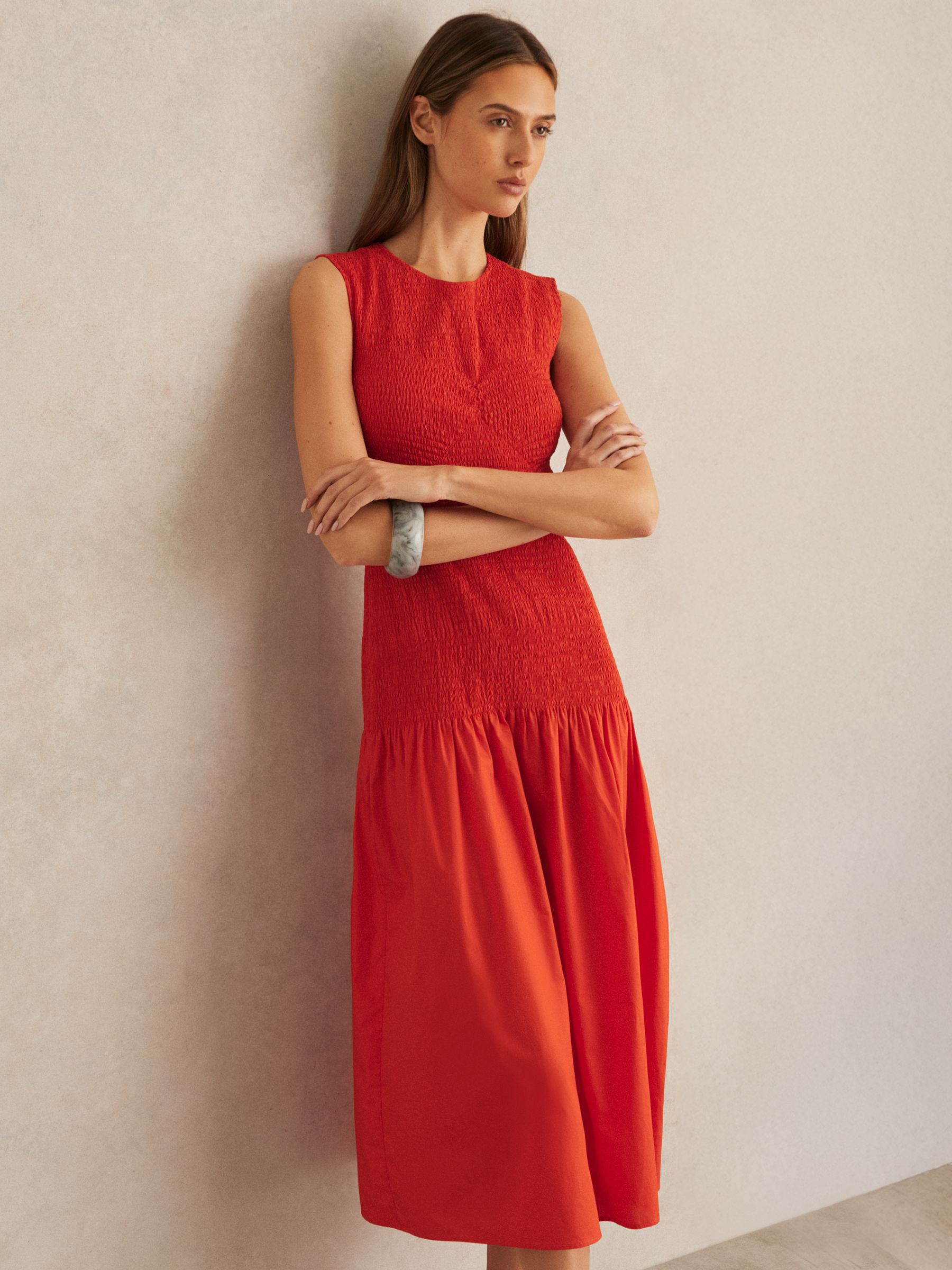 A woman wearing a sleeveless red midi dress with shirring and a flared skirt, standing against a plain wall.