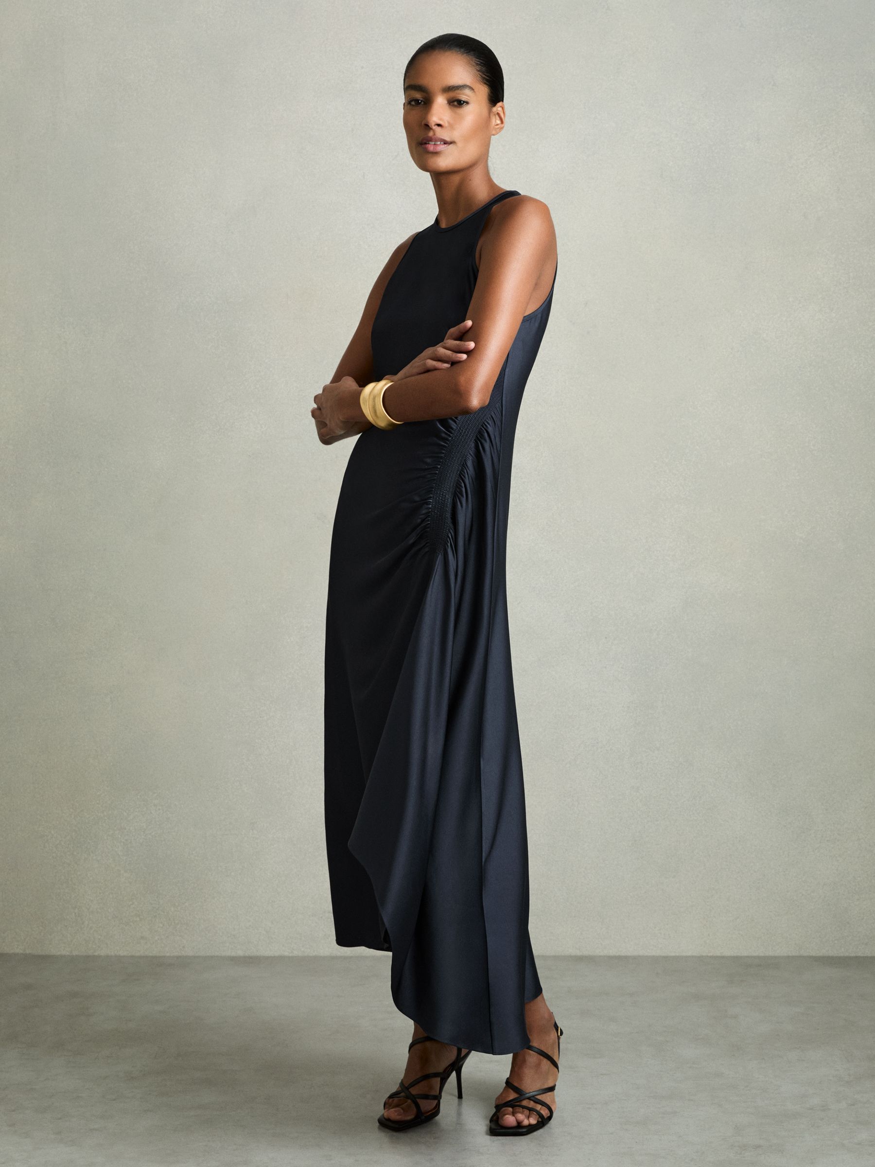 A woman wearing a navy satin midi dress with shirring on the sides, asymmetric high-low hem, round neckline, and a back button keyhole with zip.