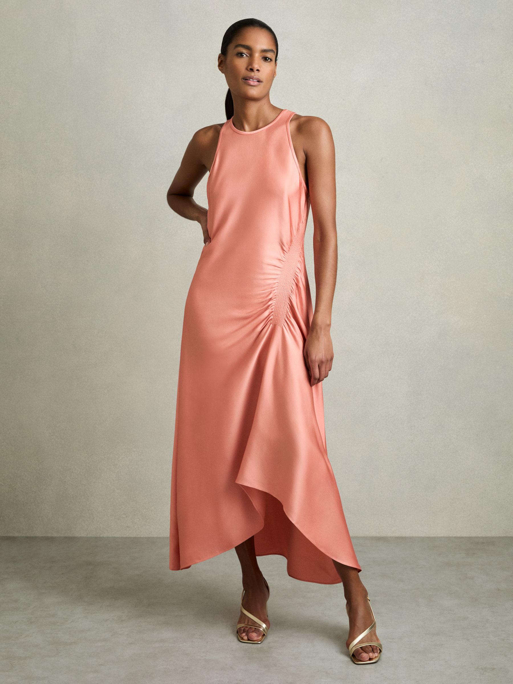 A woman wearing a coral satin midi dress with shirred side panels, round neckline, and asymmetric high-low hem.