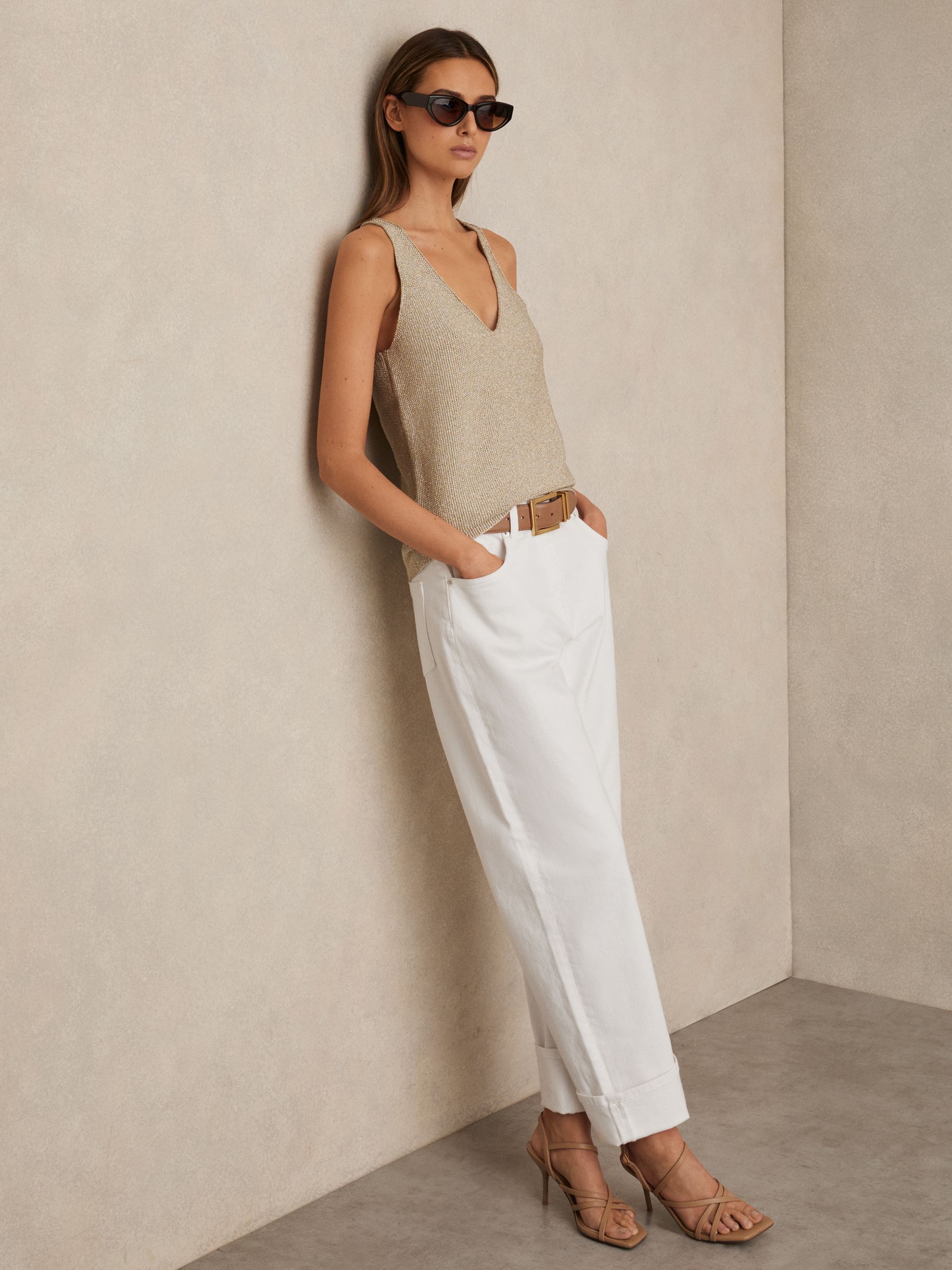 A woman wearing white petite straight leg jeans with turn-up cuffs, paired with a beige sleeveless top, sunglasses, and nude heels, standing against a beige wall.