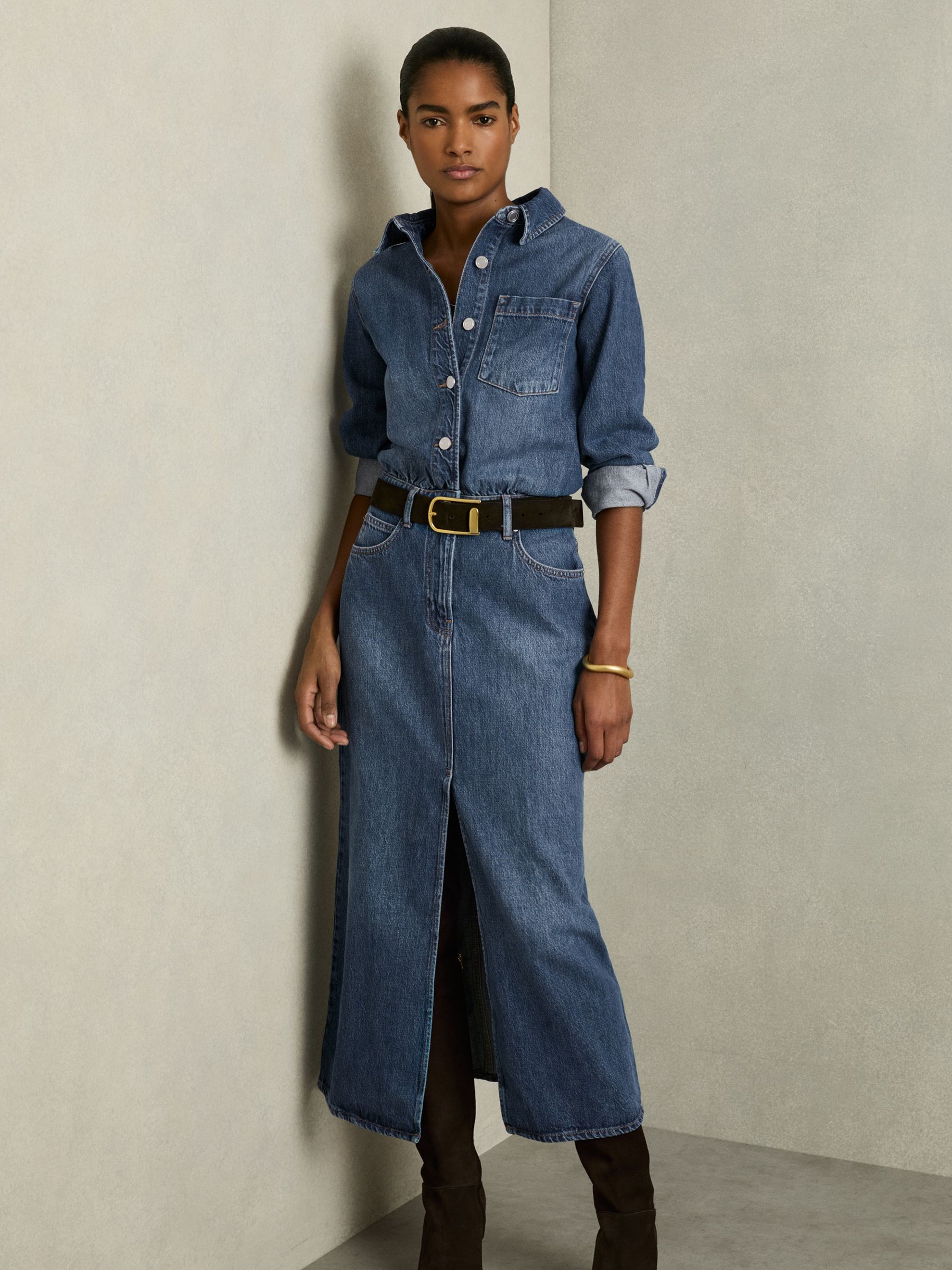 A woman wearing a blue denim midi dress with a button-through front, front slit, chest pocket, and rolled-up buttoned cuffs.