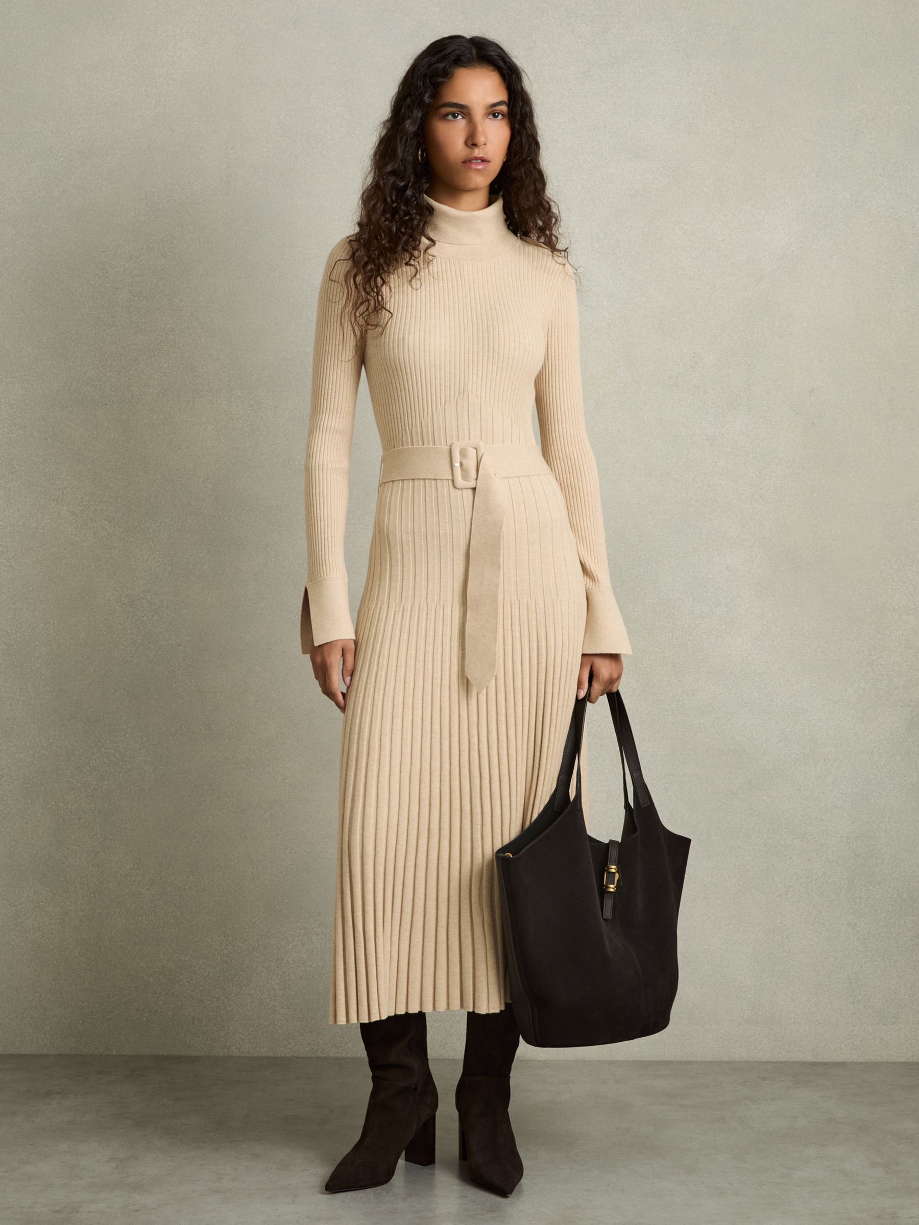 A woman wearing a beige ribbed midi dress with a roll neckline, belted waist, and slit cuffs, standing against a plain background.