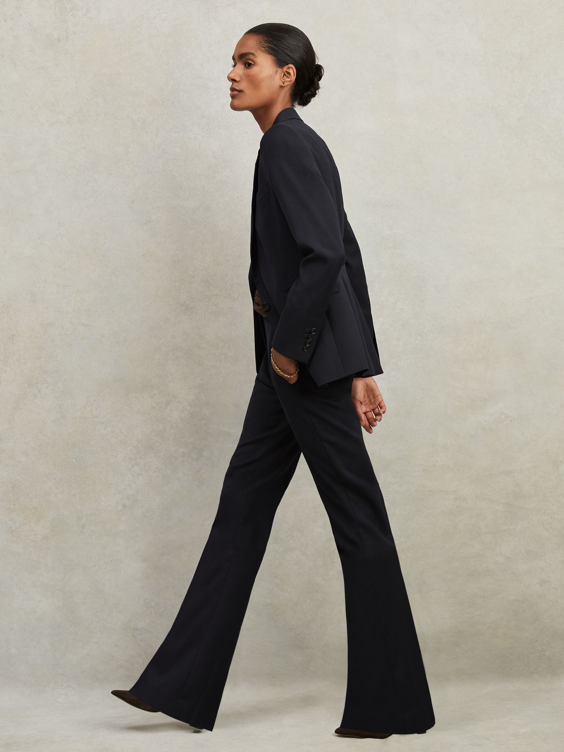 A woman wearing navy tailored flared suit trousers with a matching blazer, standing in profile against a plain background.