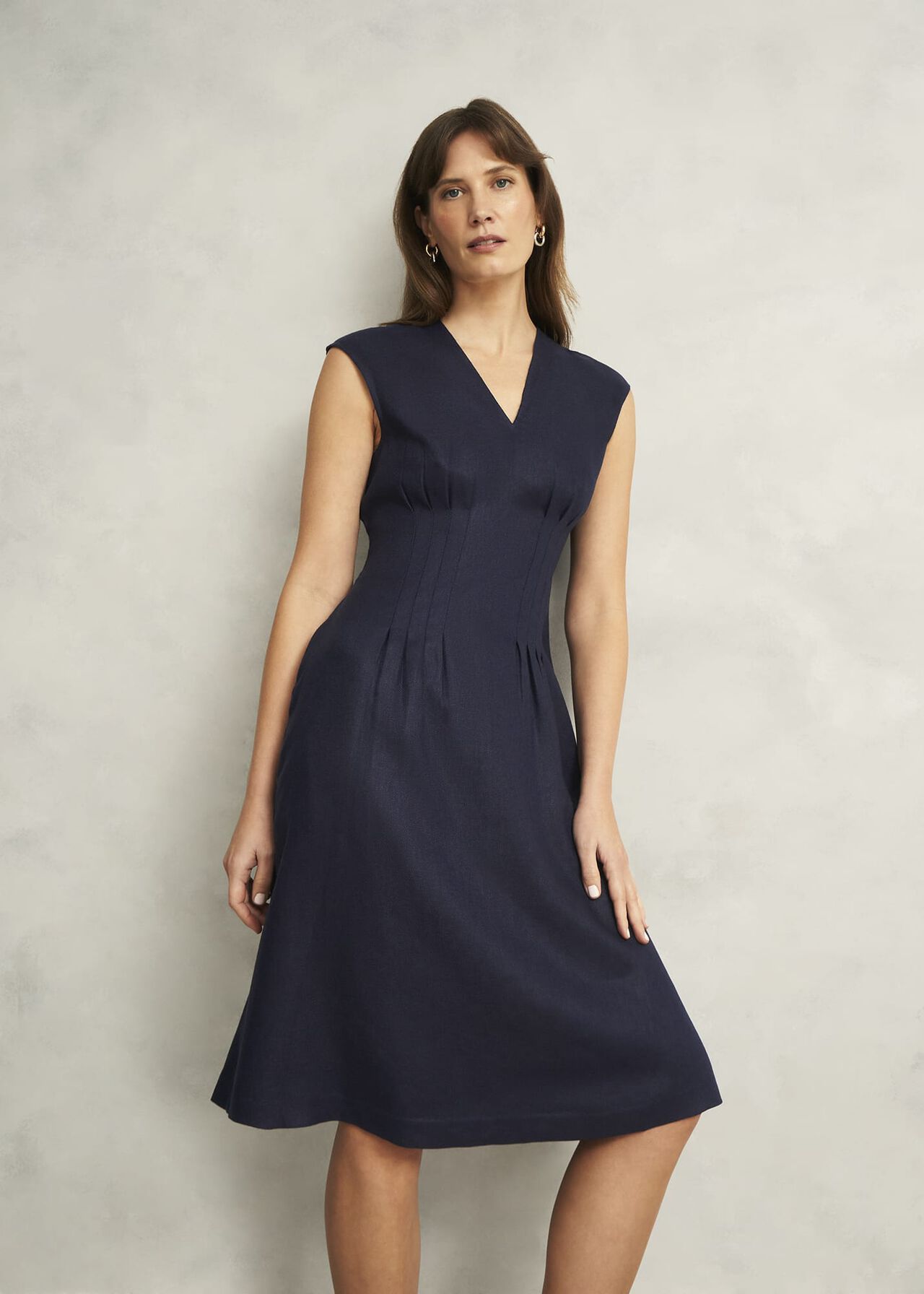 A woman wearing a beige linen dress with a V-neckline, sleeveless design, fitted waist, and midi length, standing against a plain background.